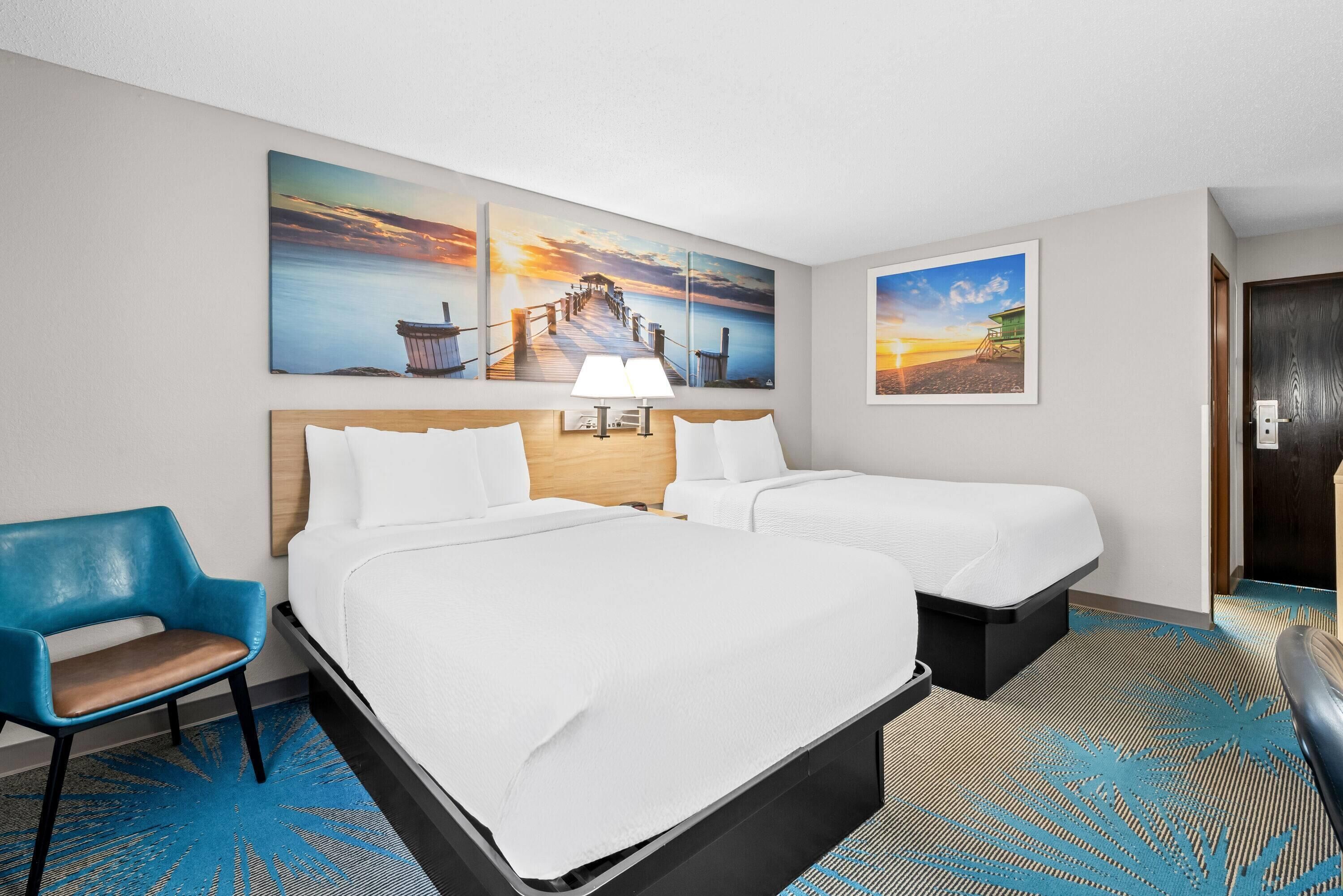 A hotel room with two beds, a desk, and a chair. There are two large wall art pieces depicting a pier and a sunset.