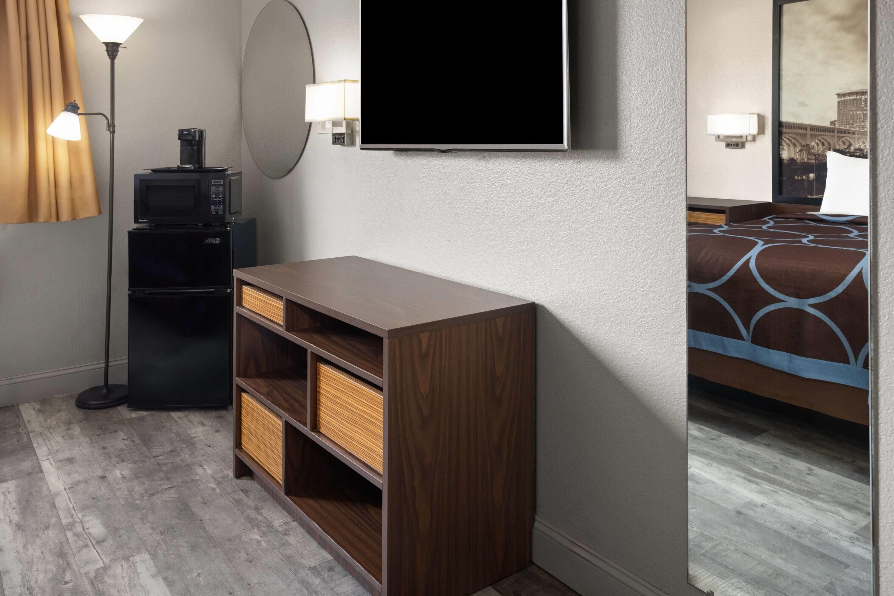 A hotel room with a flat-screen TV, a small kitchenette with a microwave, a wooden cabinet, a floor lamp, and a large mirror.