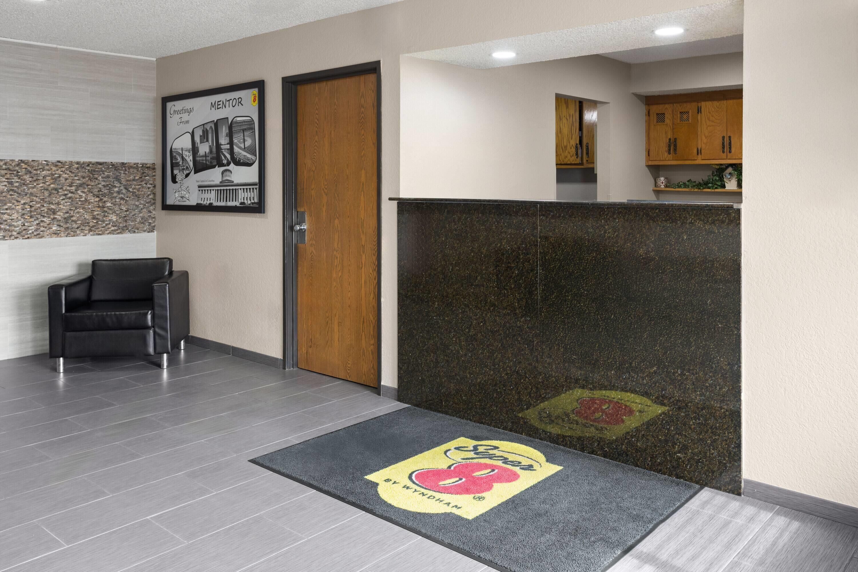 A reception area with a wooden door, a black leather chair, a reception desk, and a kitchen area in the background.