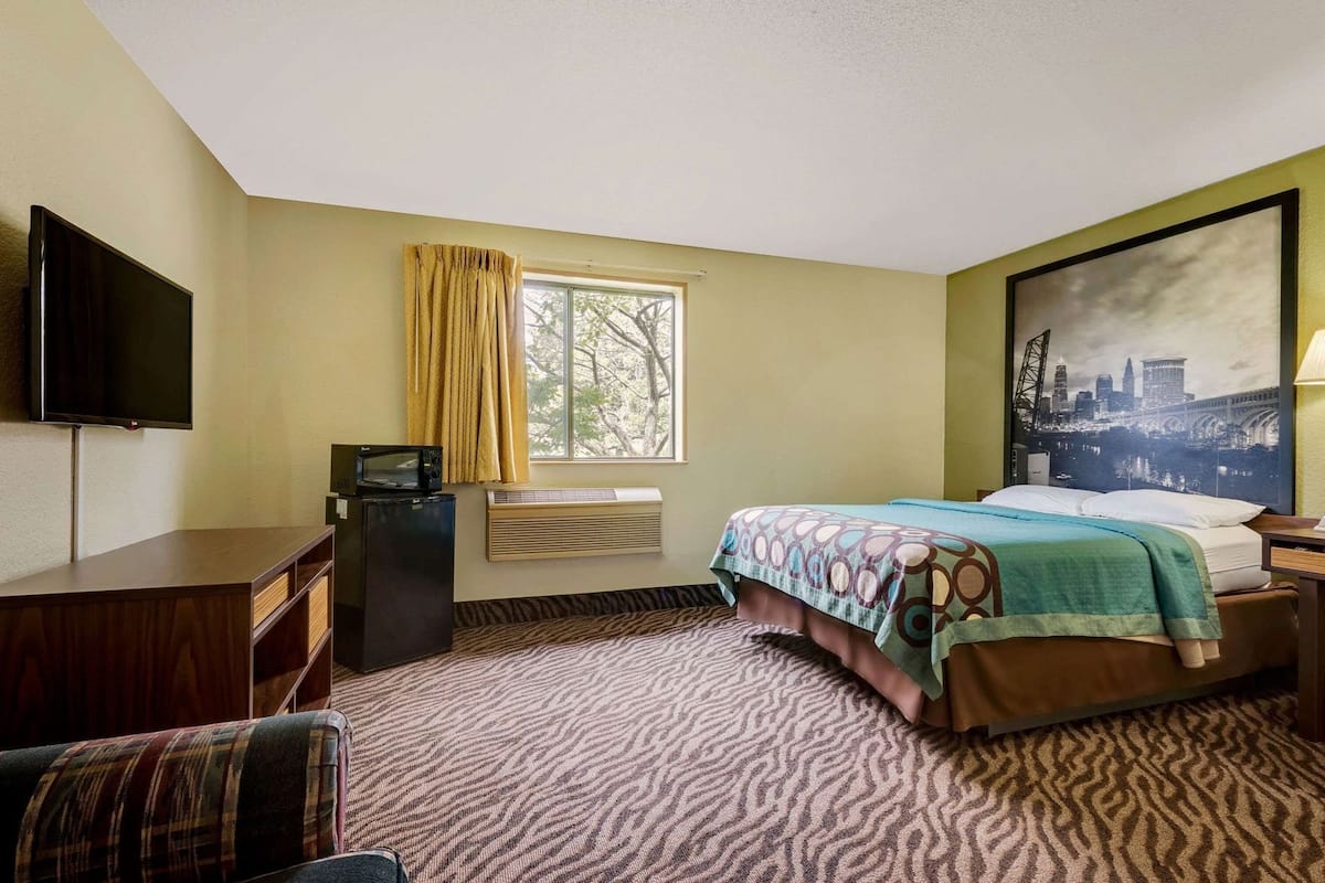 A hotel room with a bed, a desk with a flat-screen TV, a microwave, a window with curtains, an air conditioning unit, and a cityscape painting.