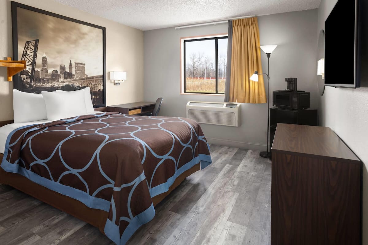A hotel room with a bed, desk, air conditioning unit, lamp, and a cityscape painting.