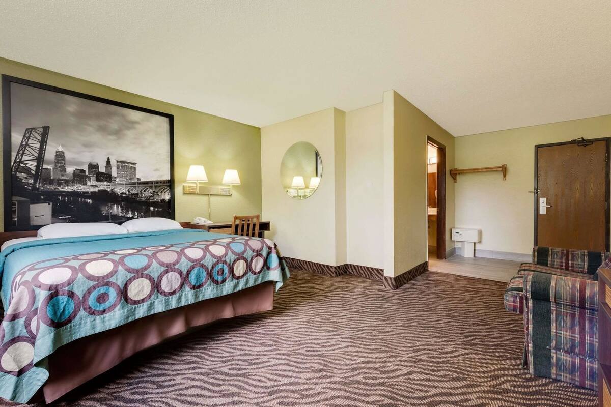 A hotel room with a bed, two chairs, a desk, a mirror, and a cityscape painting on the wall.
