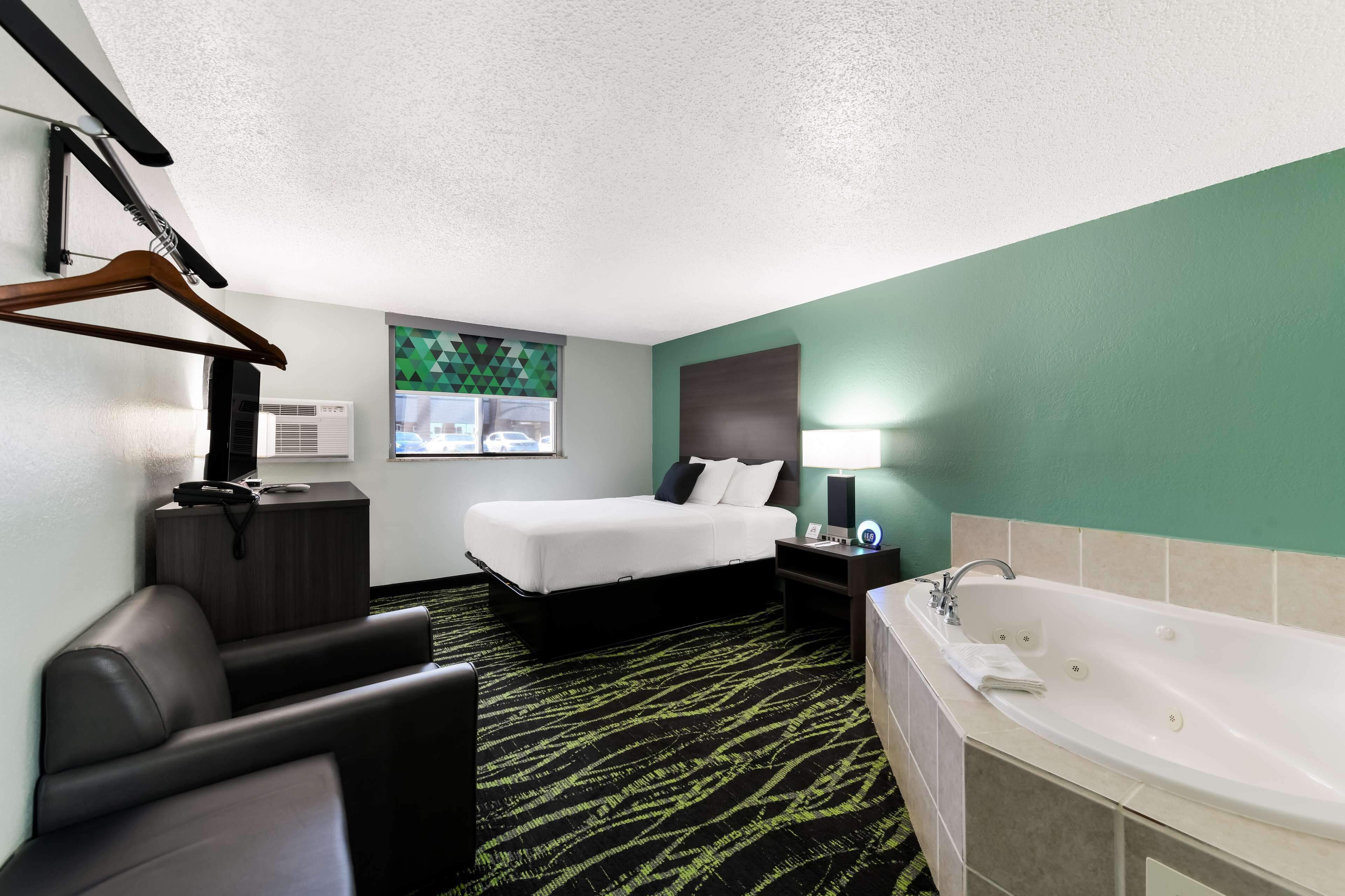 A hotel room with a bed, a desk, a chair, a desk lamp, a wall-mounted TV, a window with a view of cars, and a bathtub.