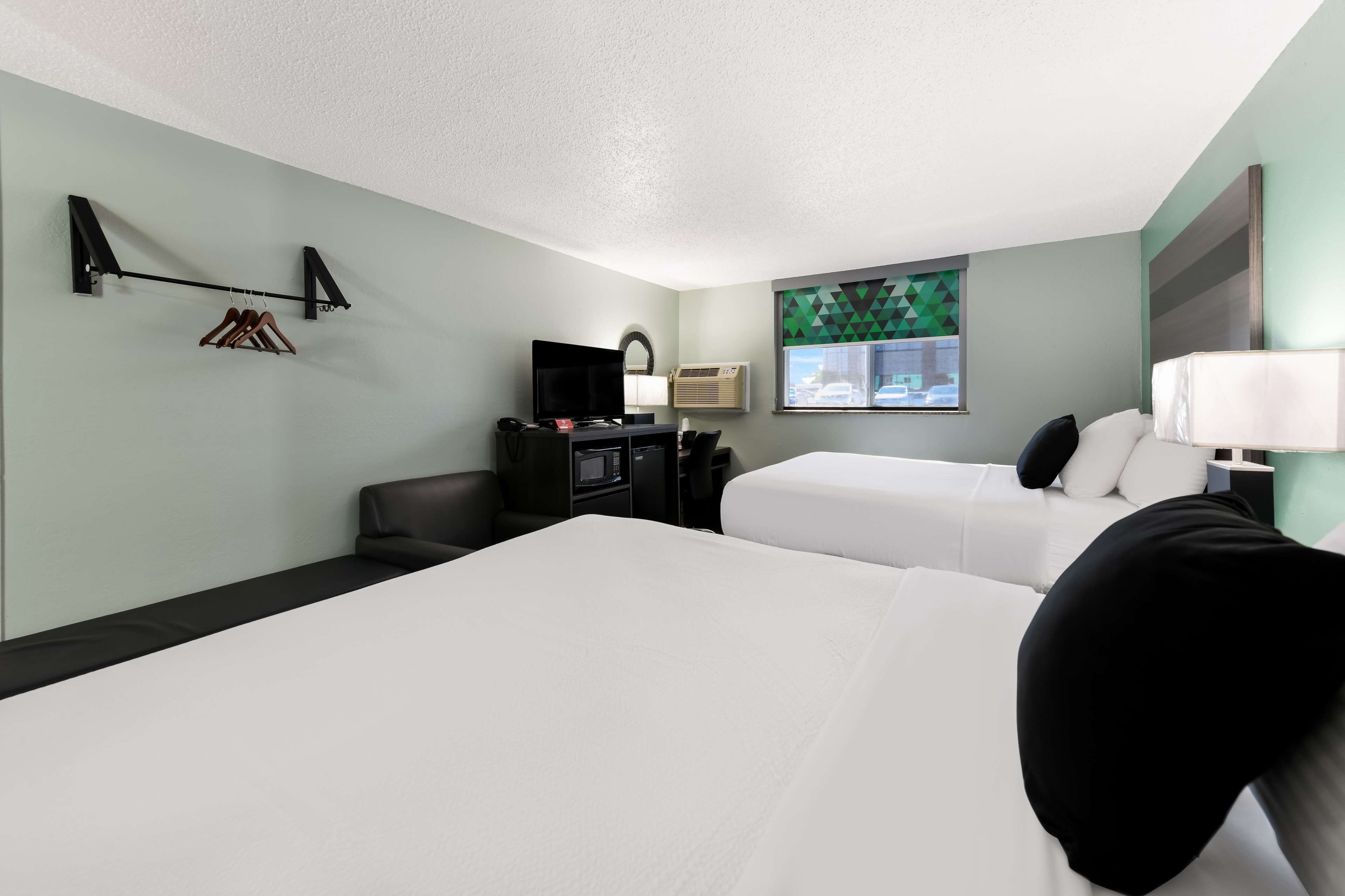 Une chambre d’hôtel avec deux lits, un bureau avec un ordinateur, une télévision et une fenêtre donnant sur un paysage aux formes géométriques.