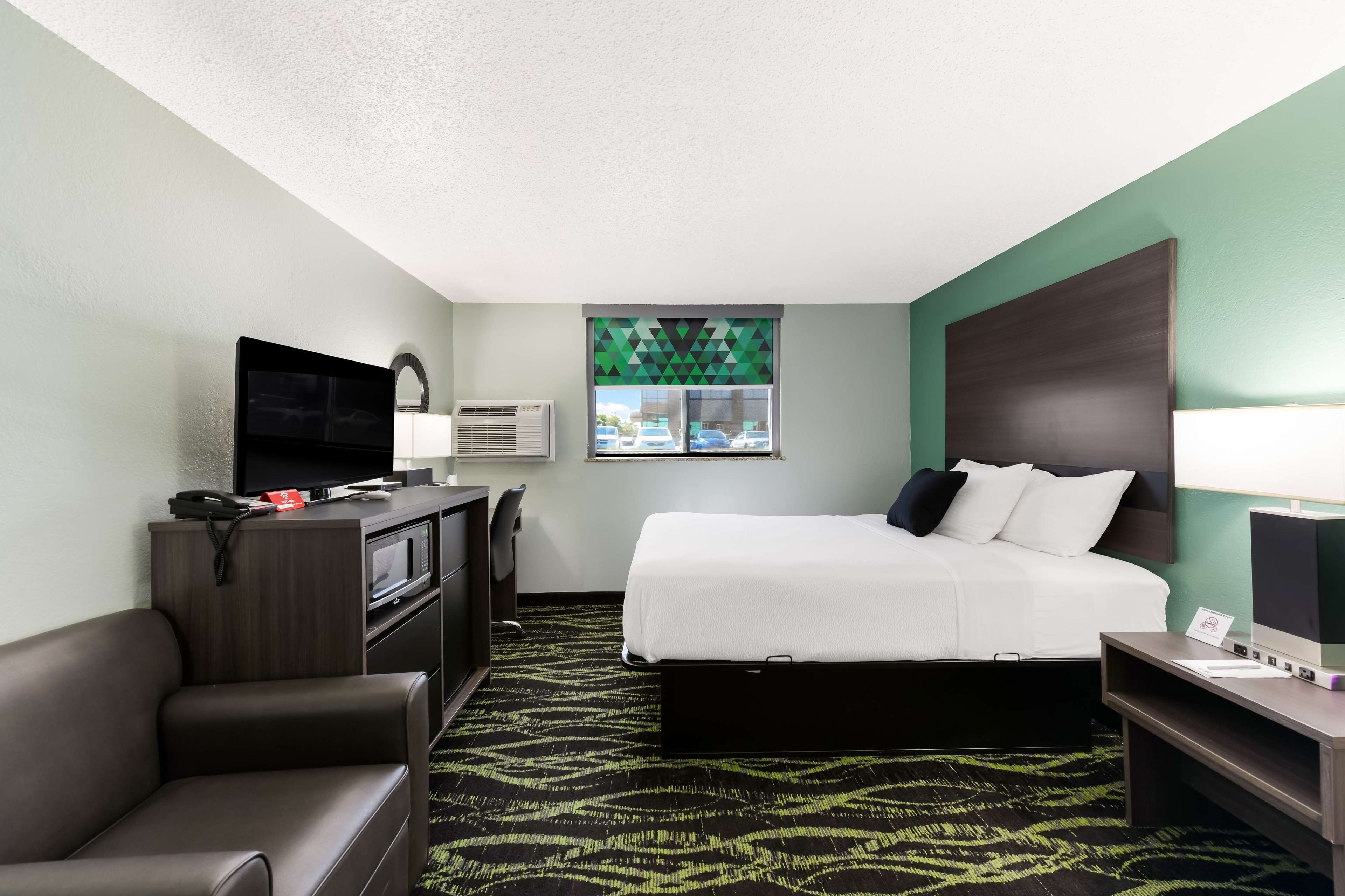 A hotel room with a bed, a desk, a TV, a chair, and a window with a view of cars.
