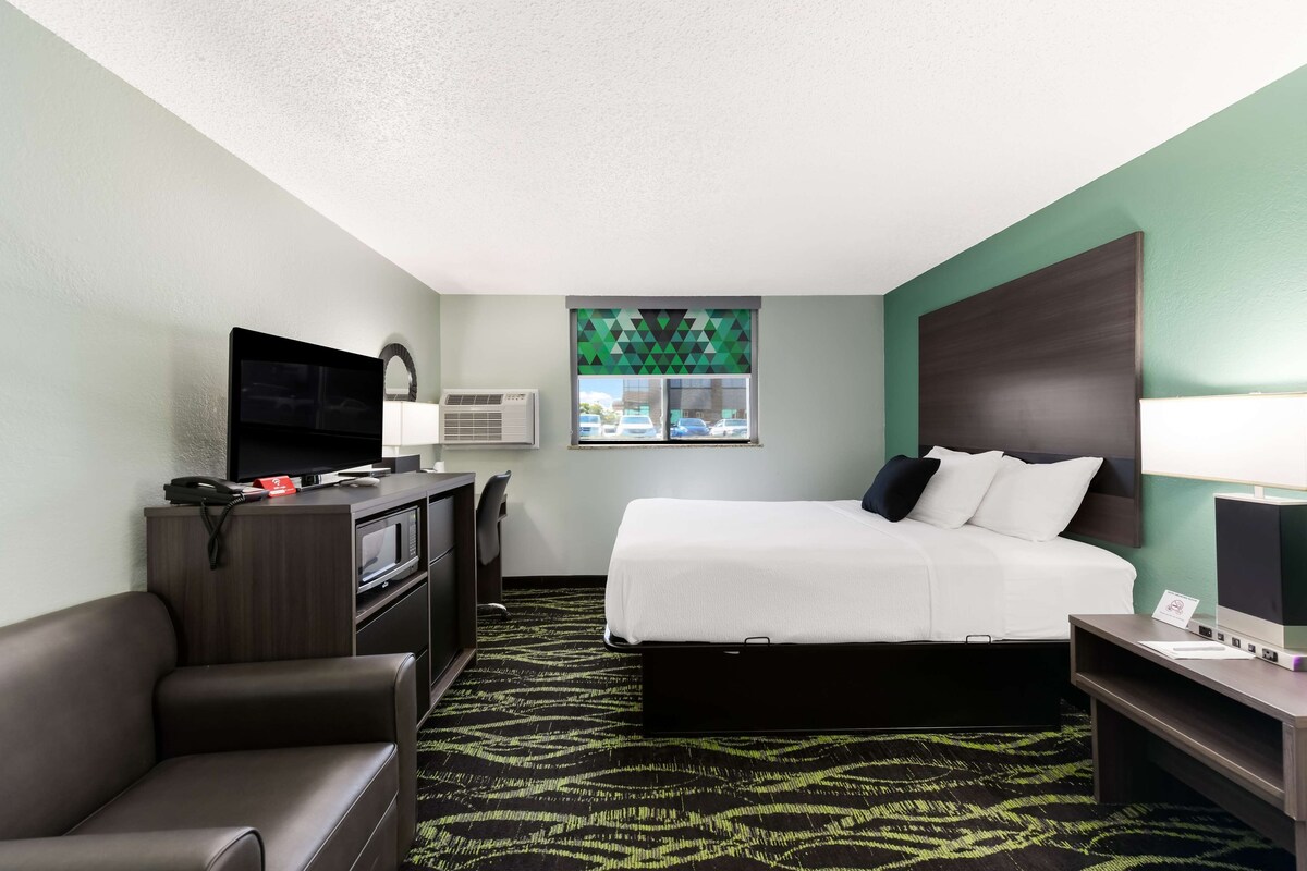 A hotel room with a bed, a desk, a TV, a chair, and a window with a view of cars.