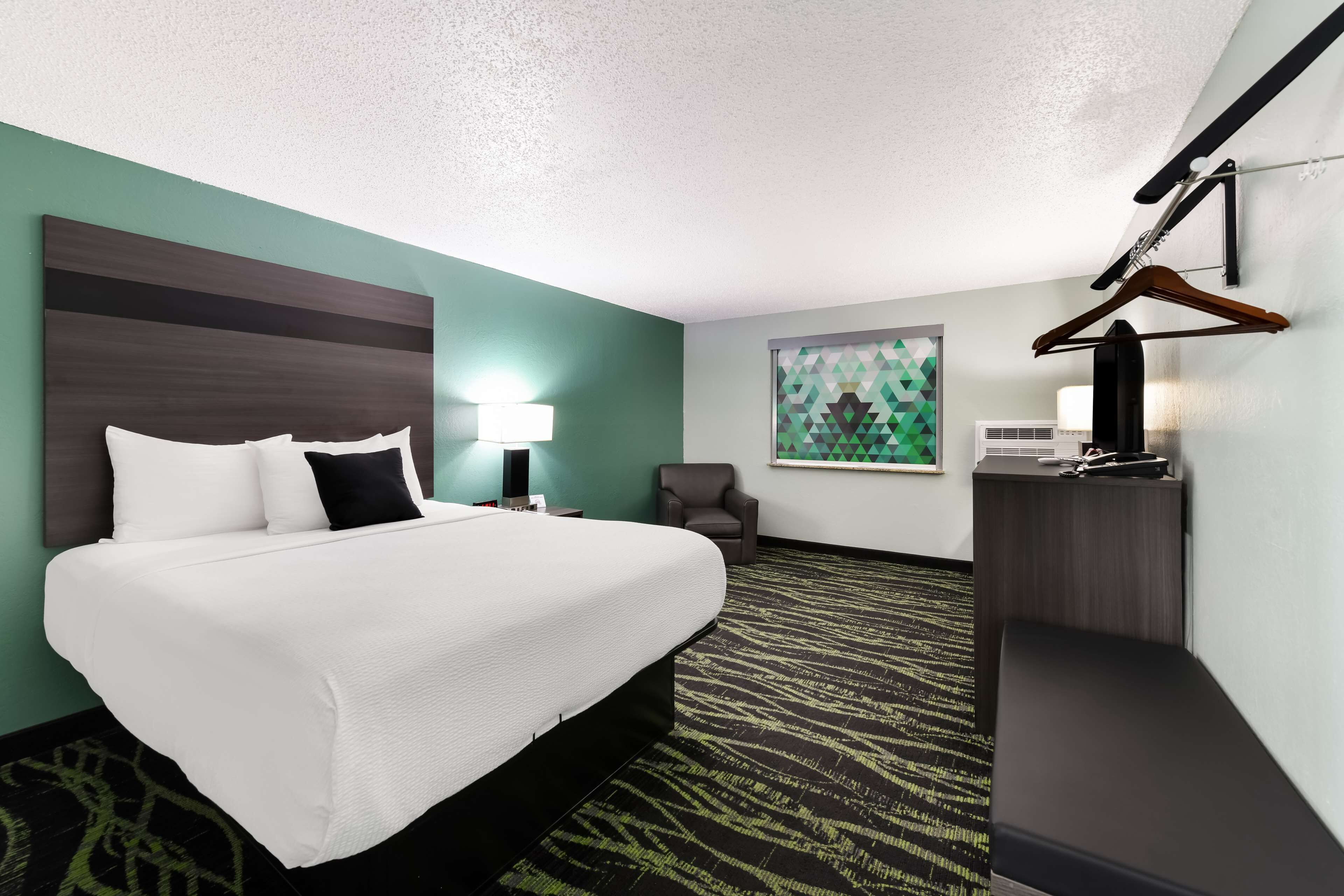 A hotel room with a bed, a desk, a chair, a painting, and a coat rack.