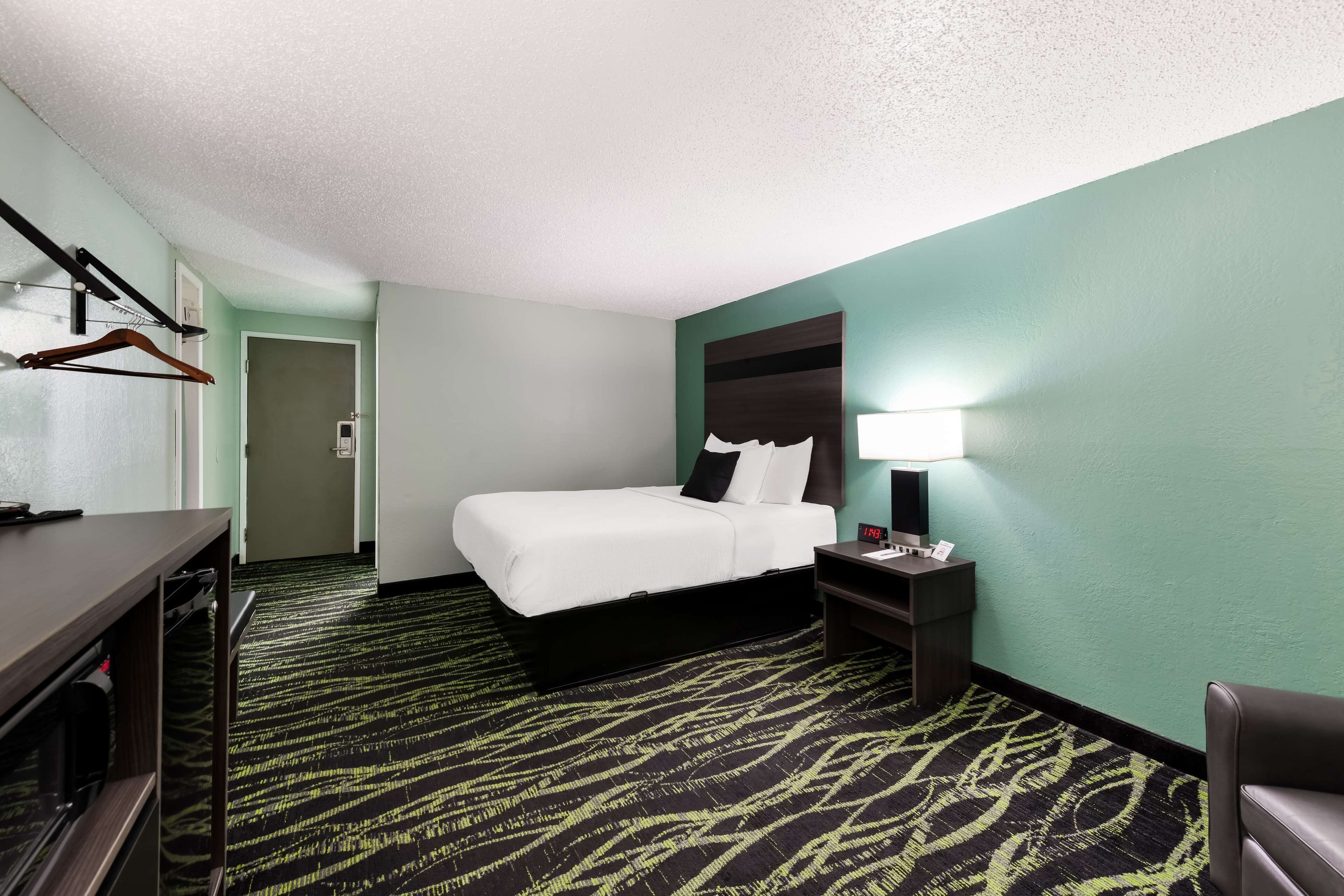 A hotel room with a bed, a desk, a chair, a nightstand, and a lamp.
