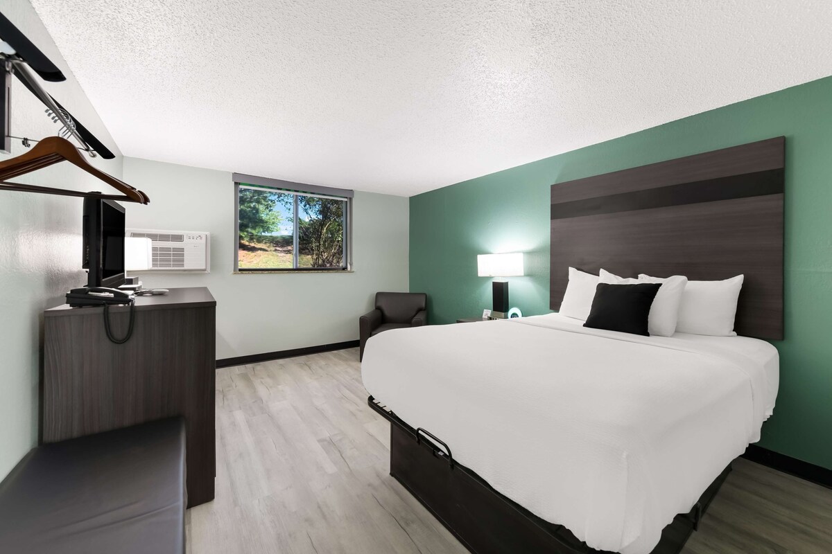 A hotel room with a bed, desk, chair, and a window with a view of greenery.