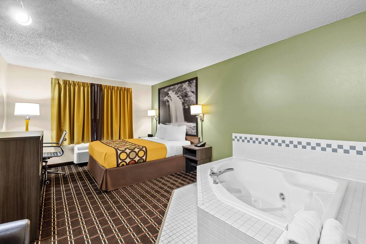 A hotel room with a bed, a desk, a chair, a desk lamp, a nightstand, a telephone, a bathroom with a bathtub, and a shower.
