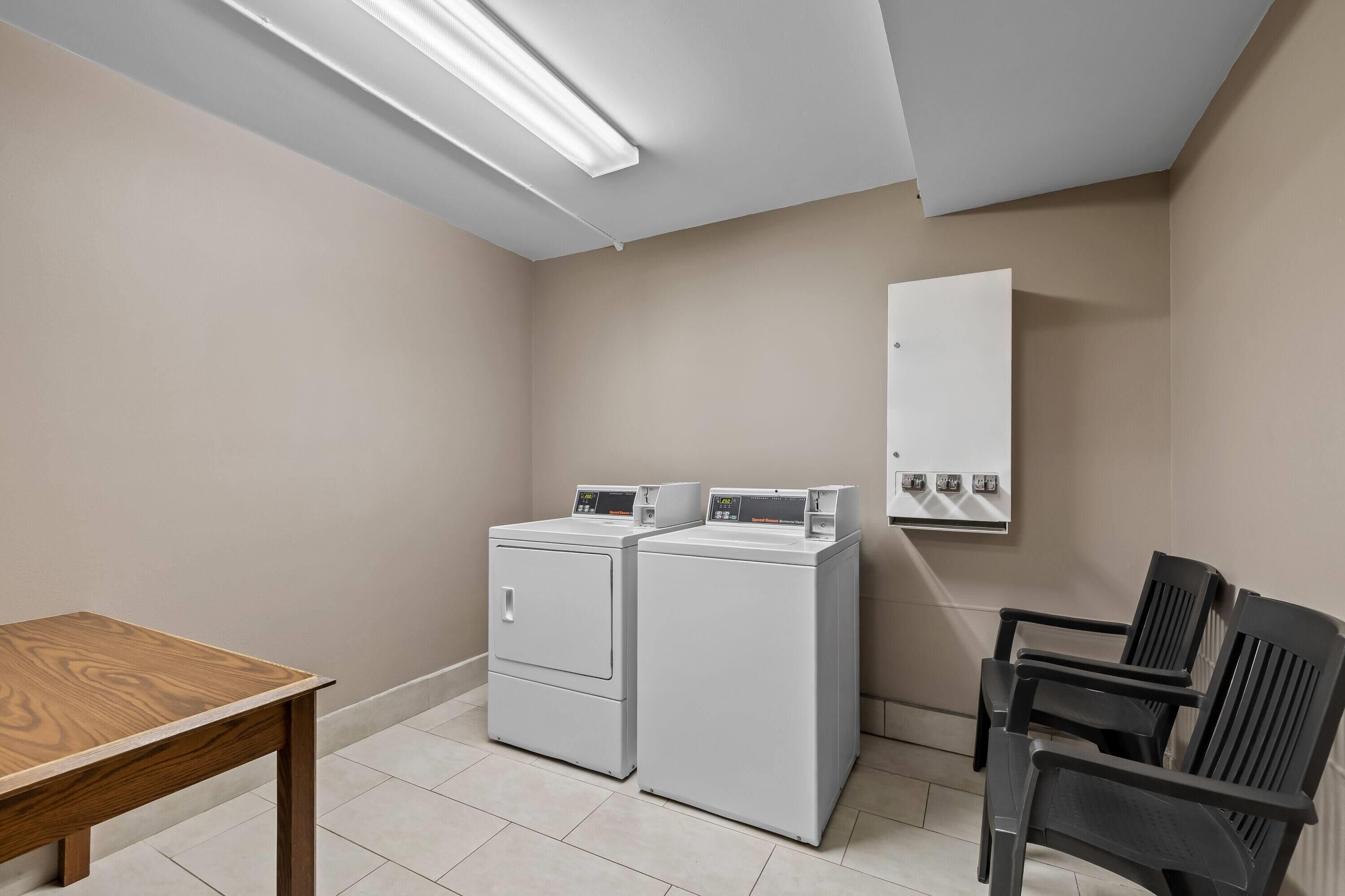 Una sala de bugaderia amb dues rentadores i assecadores blanques, una taula de fusta i cadires negres.