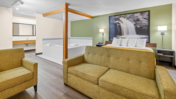 Habitación de hotel con una cama grande, dos sillones, un baño con bañera y un cuadro grande en la pared que representa una cascada.