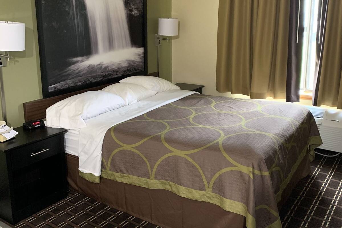 A hotel room with a bed, a nightstand, a lamp, a large wall painting, a window with curtains, and a floor with a checkered pattern.
