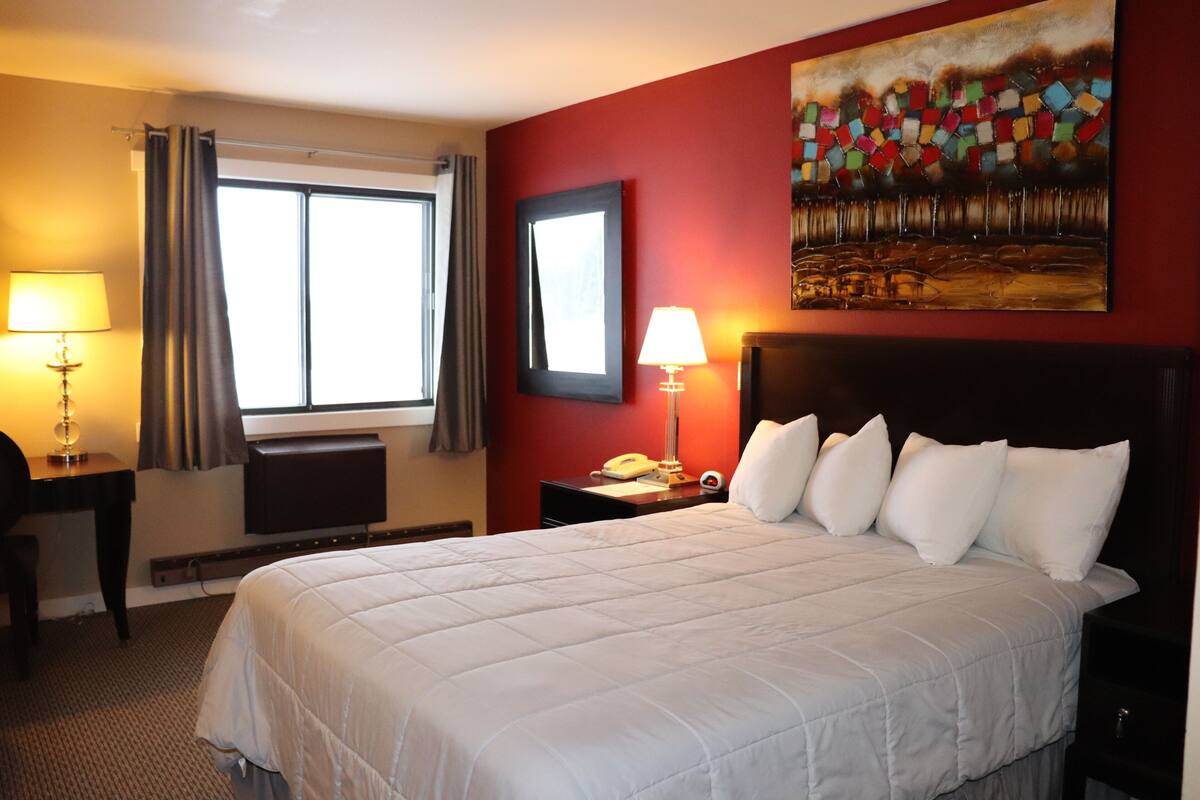 Une chambre d’hôtel avec un grand lit, un bureau avec une lampe, une fenêtre avec des rideaux, un tableau au mur et une table de chevet avec une lampe et un téléphone.
