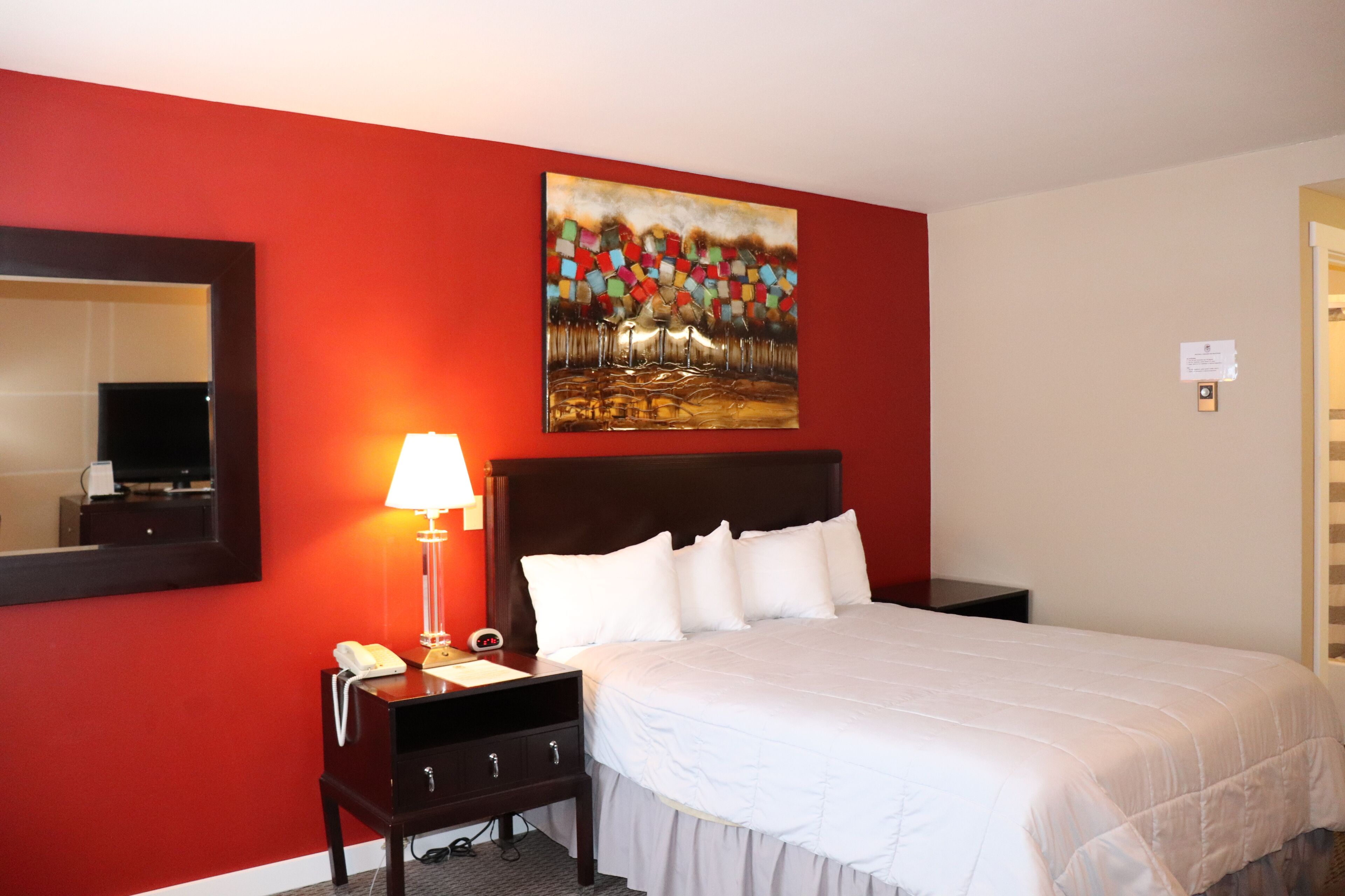 A hotel room with a red wall, a bed with white bedding, a nightstand with a lamp, and a painting above the bed.