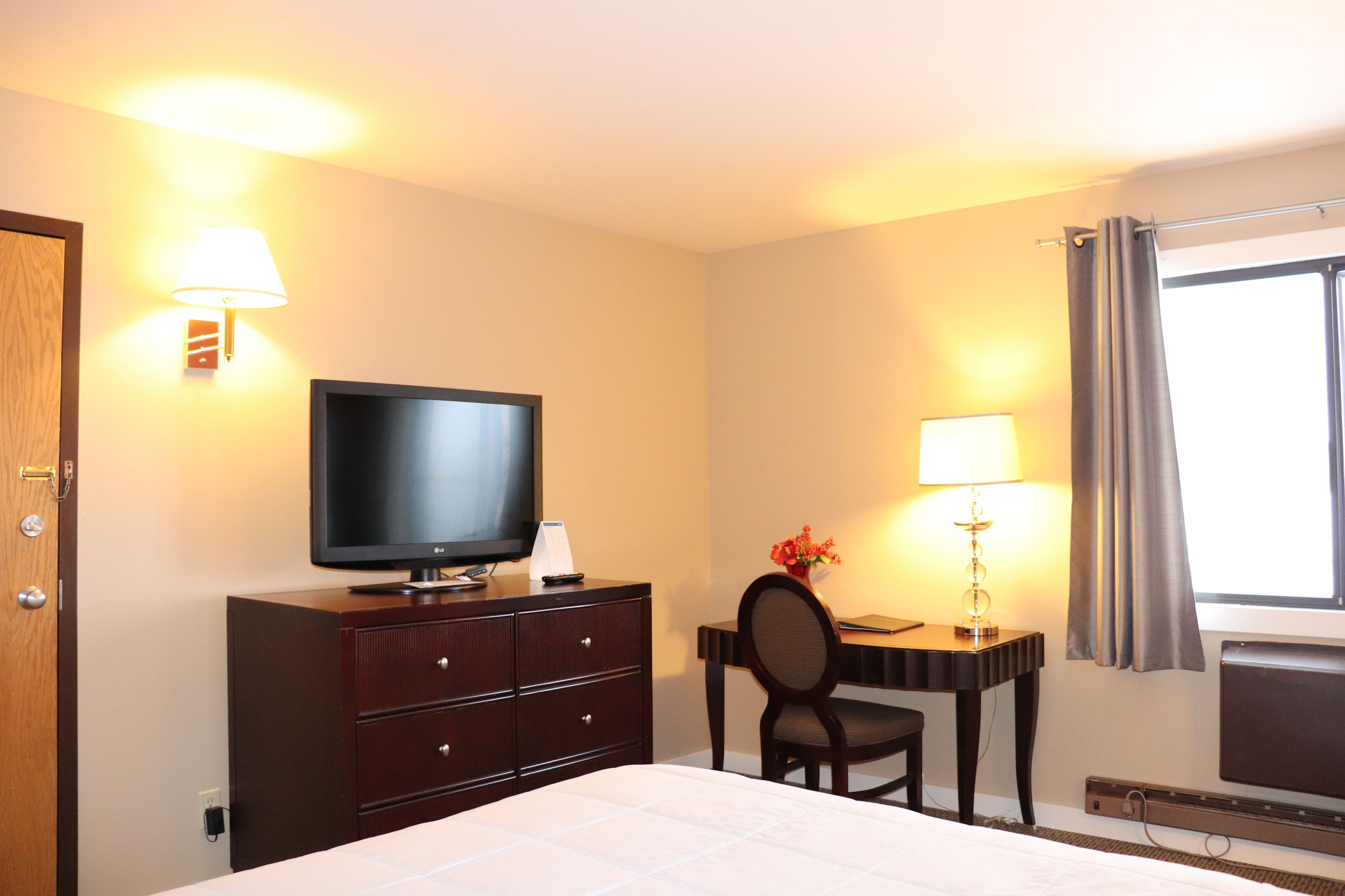Ein Hotelzimmer mit Bett, Fernseher, Schreibtisch mit Lampe und einem Fenster mit Vorhängen.