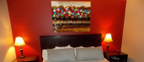 Una cama bien hecha con ropa blanca, dos mesitas de noche con lámparas y un cuadro abstracto colorido en la pared.