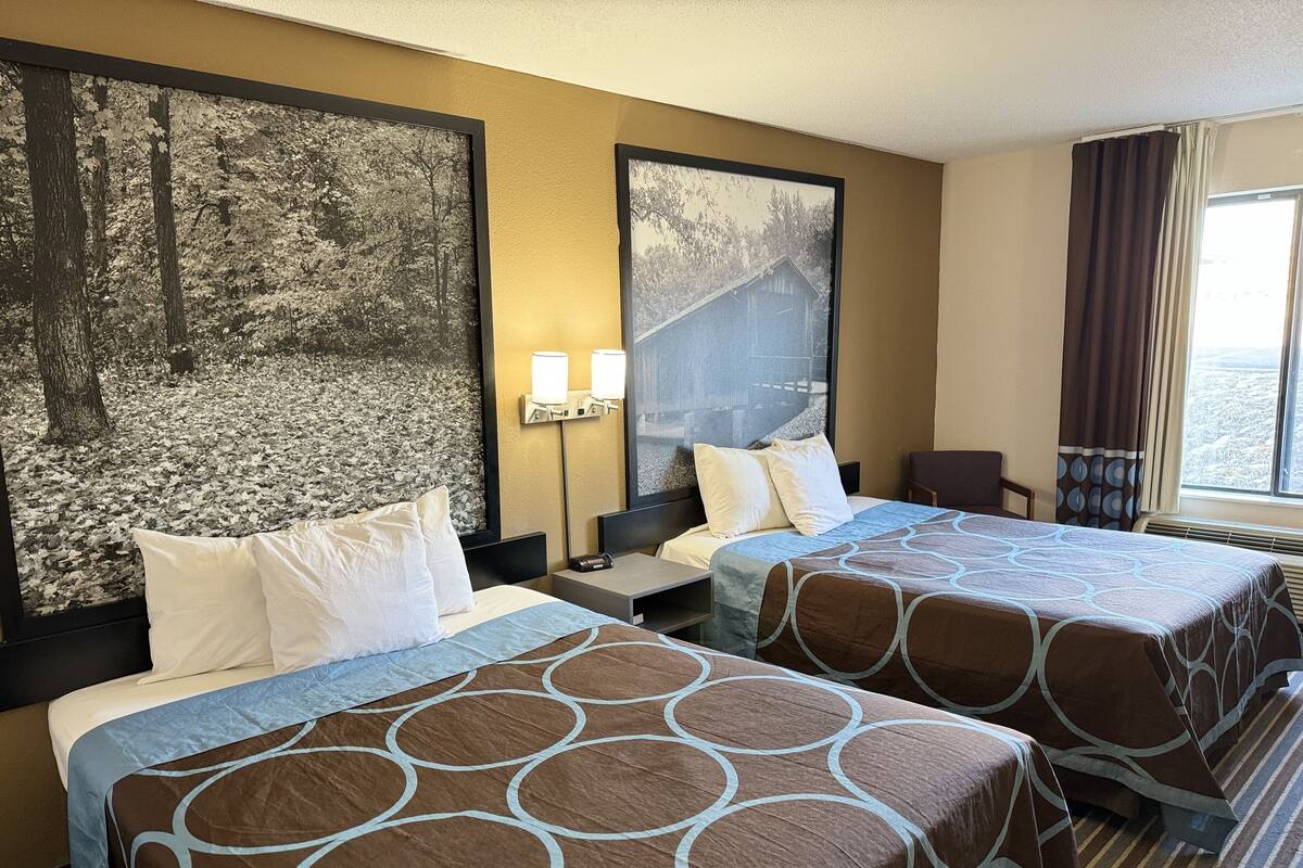 Ein Hotelzimmer mit zwei Betten, einem großen gerahmten Bild von einem Wald, einem Fenster mit Vorhängen und einem Sessel.