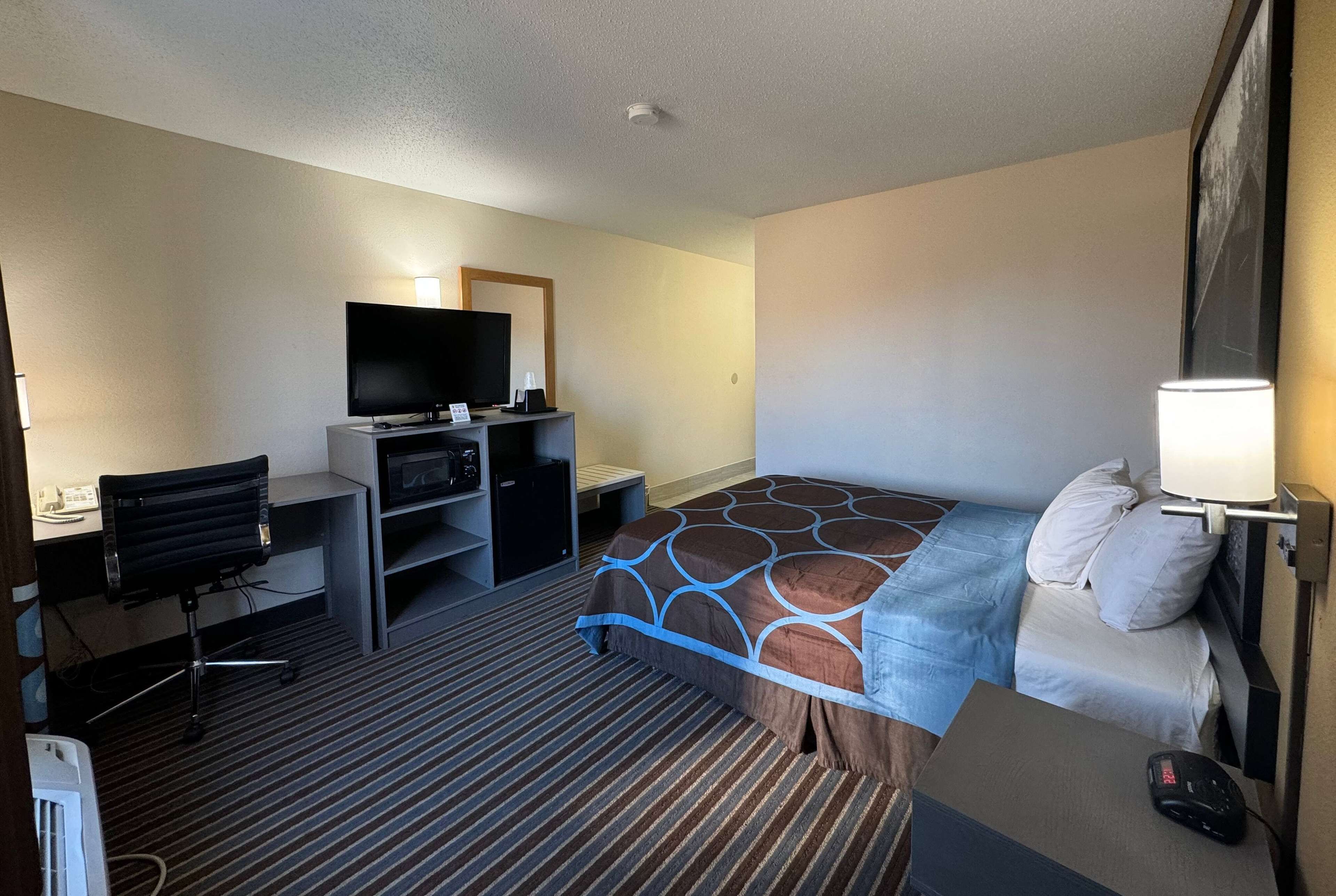 Quarto de hotel com cama, escrivaninha, computador, cadeira, televisão e luminária.