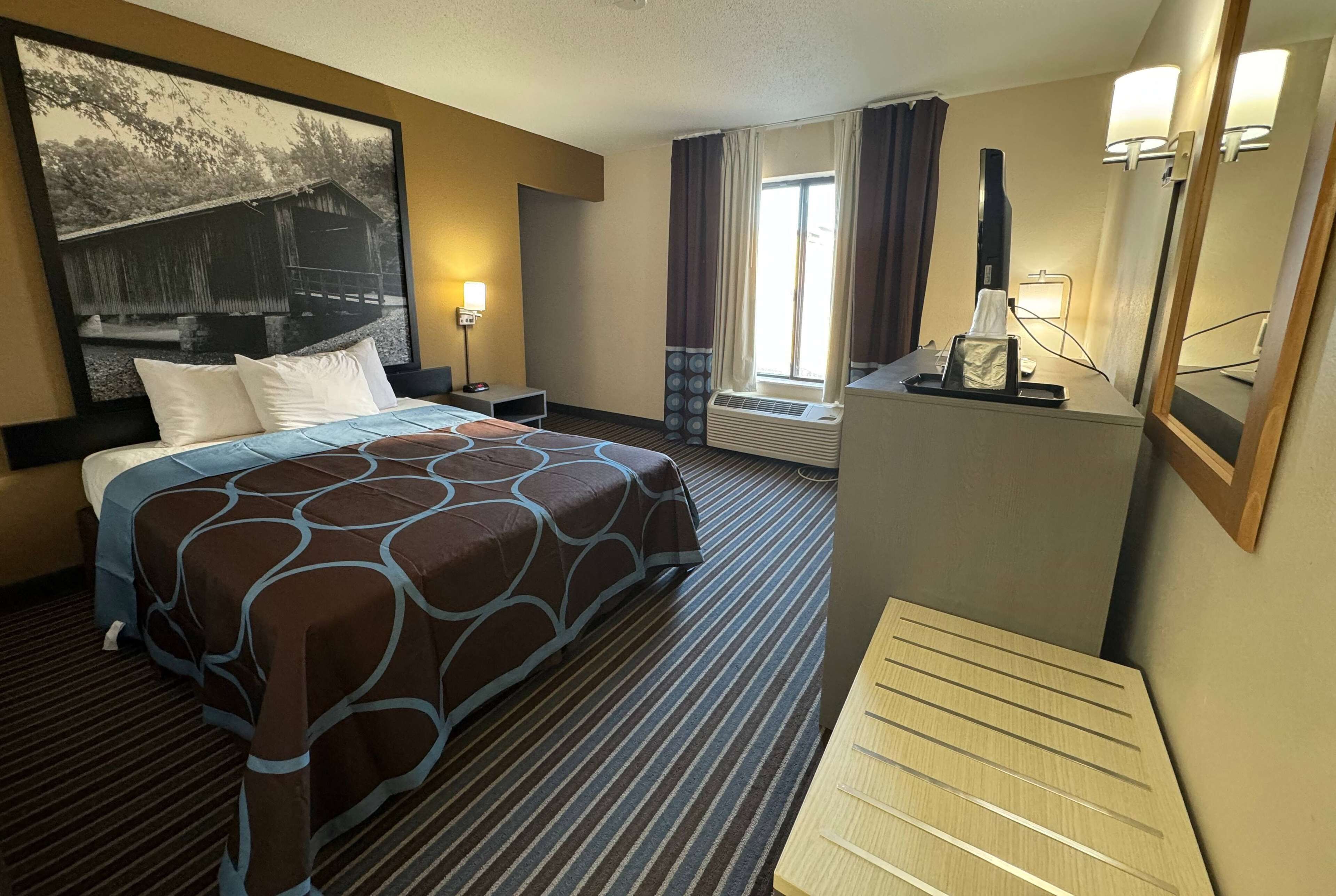 A hotel room with a large bed, a desk with a lamp, a window with curtains, and a large painting on the wall.