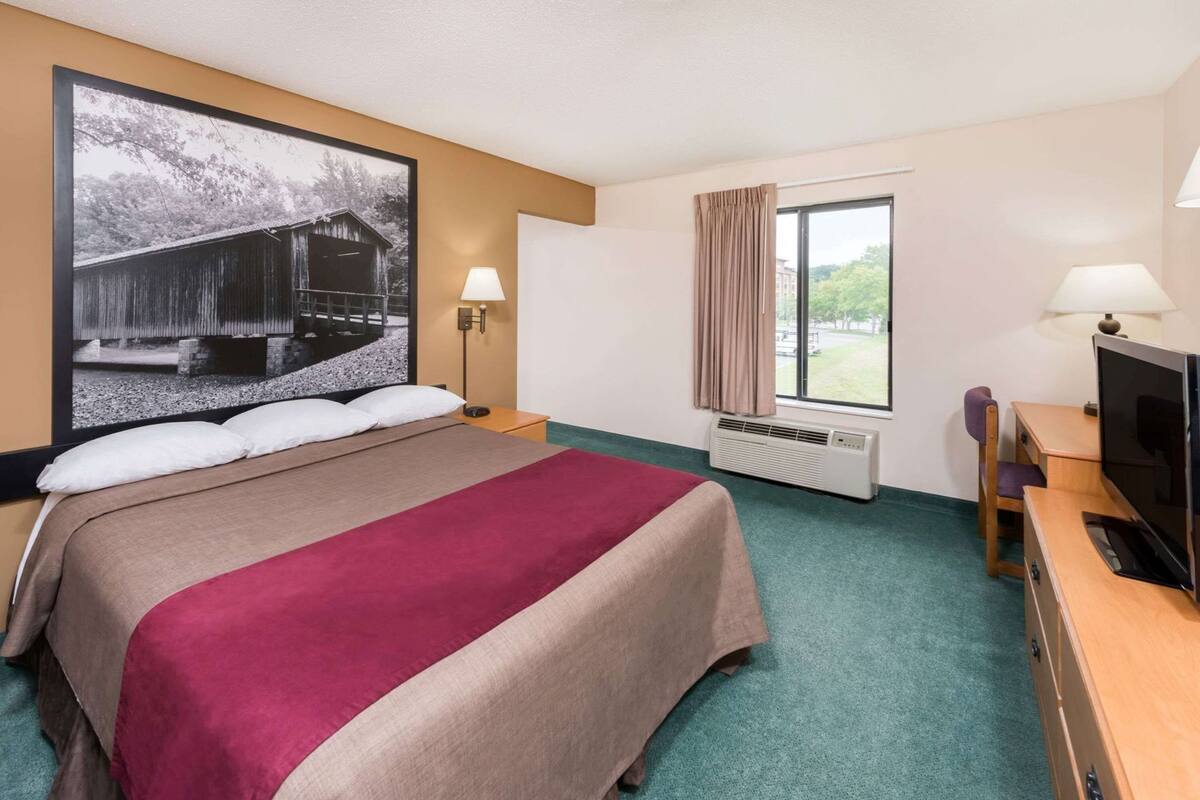 Ein Hotelzimmer mit einem Bett, einem Schreibtisch mit Fernseher, einem Fenster mit Vorhängen, einer Wand-Klimaanlage und einem großen, gerahmten Bild.