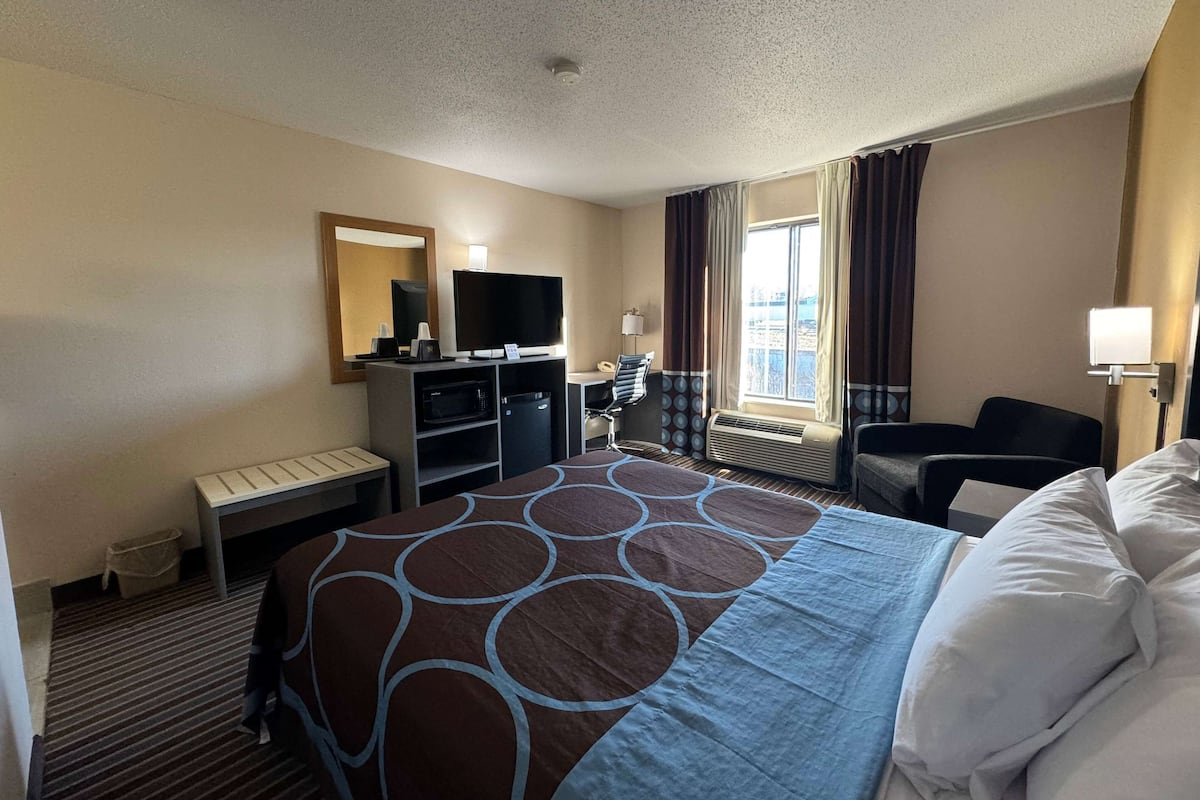 Ein Hotelzimmer mit einem großen Bett, einem Schreibtisch mit Flachbildfernseher, einem Sessel, einem Fenster mit Vorhängen und einer Klimaanlage.