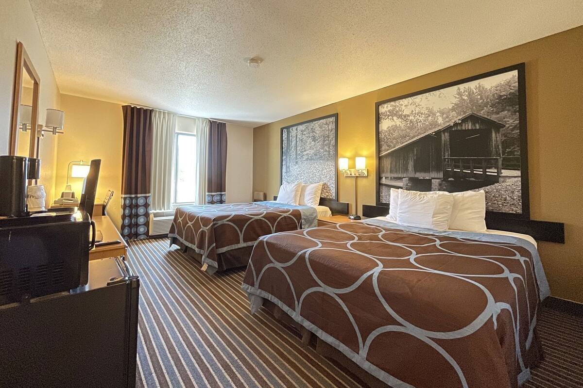 Ein Hotelzimmer mit zwei Betten, einem Schreibtisch mit Stuhl, einer Kaffeemaschine und einem großen Fenster mit Vorhängen.