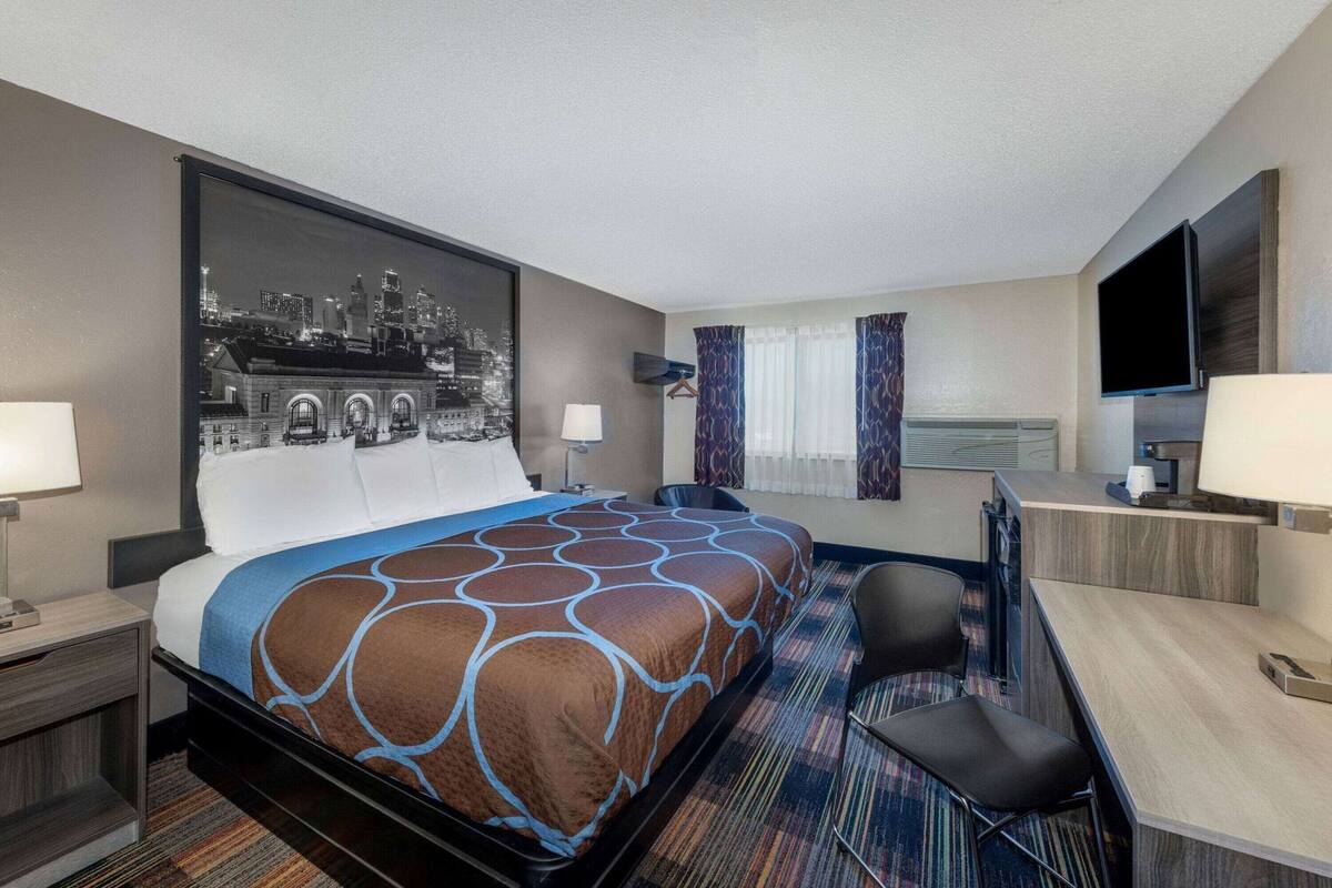 Une chambre d’hôtel comprenant un lit, un bureau, une chaise, un téléviseur et une œuvre d’art représentant un paysage urbain accrochée au mur.