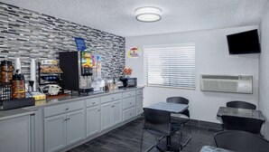 A modern kitchen with grey cabinets, a coffee station, a breakfast sign, and a TV mounted on the wall. - Super 8 by Wyndham Kansas City at Barry Road/Airport (Kansas City)