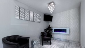 A modern living room with a black leather sofa, a round metallic side table, a dark leather armchair, a flat-screen TV mounted on the wall, and a decorative chandelier. - Super 8 by Wyndham Kansas City at Barry Road/Airport (Kansas City)