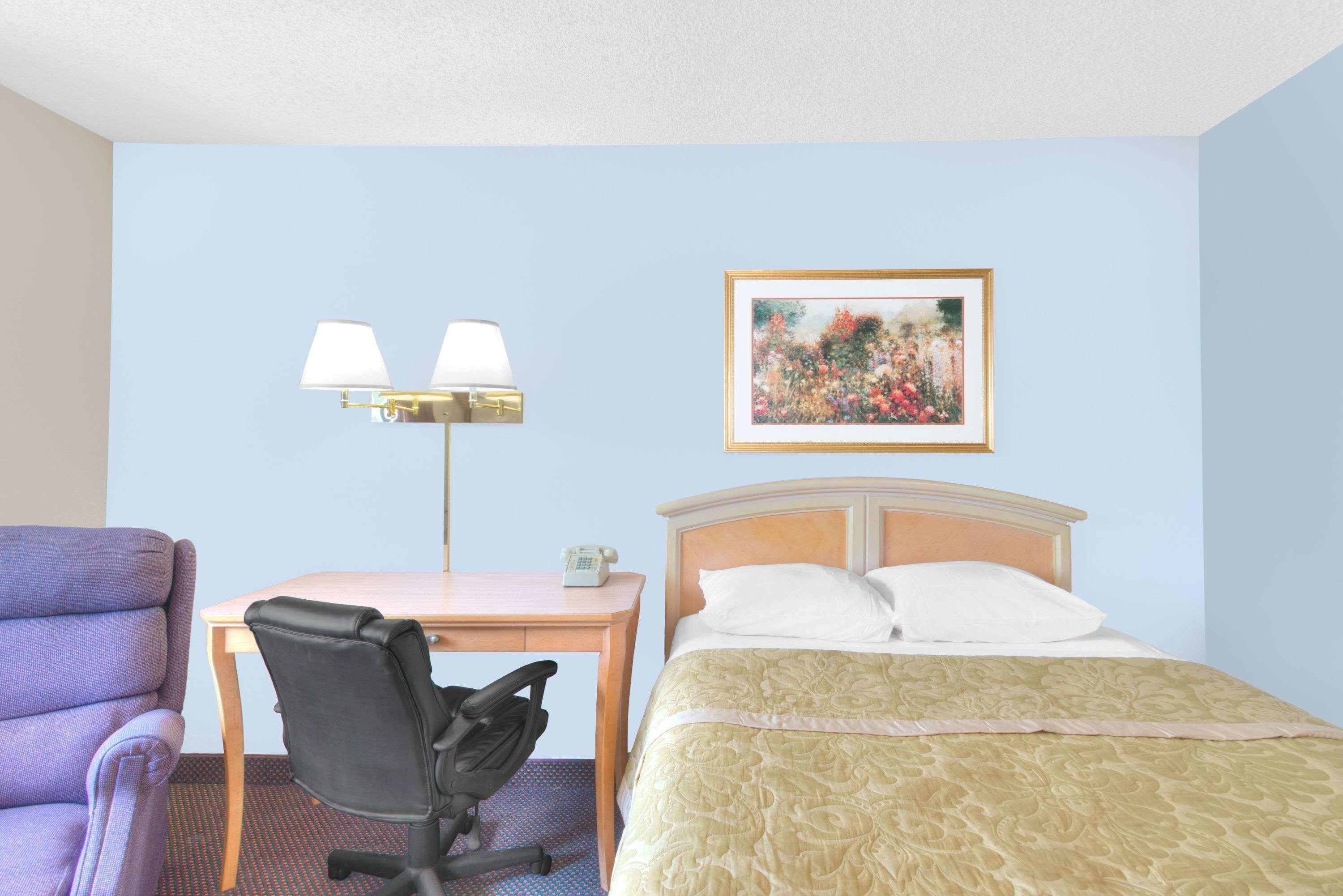 A hotel room with a bed, a desk with a chair, a lamp, and a framed picture on the wall.