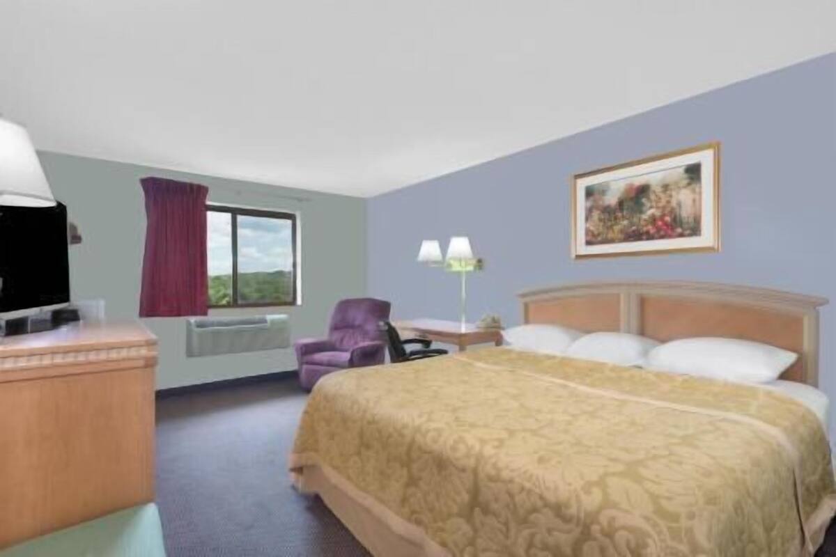 A hotel room with a bed, a desk, a chair, a window with curtains, a lamp, and a painting on the wall.