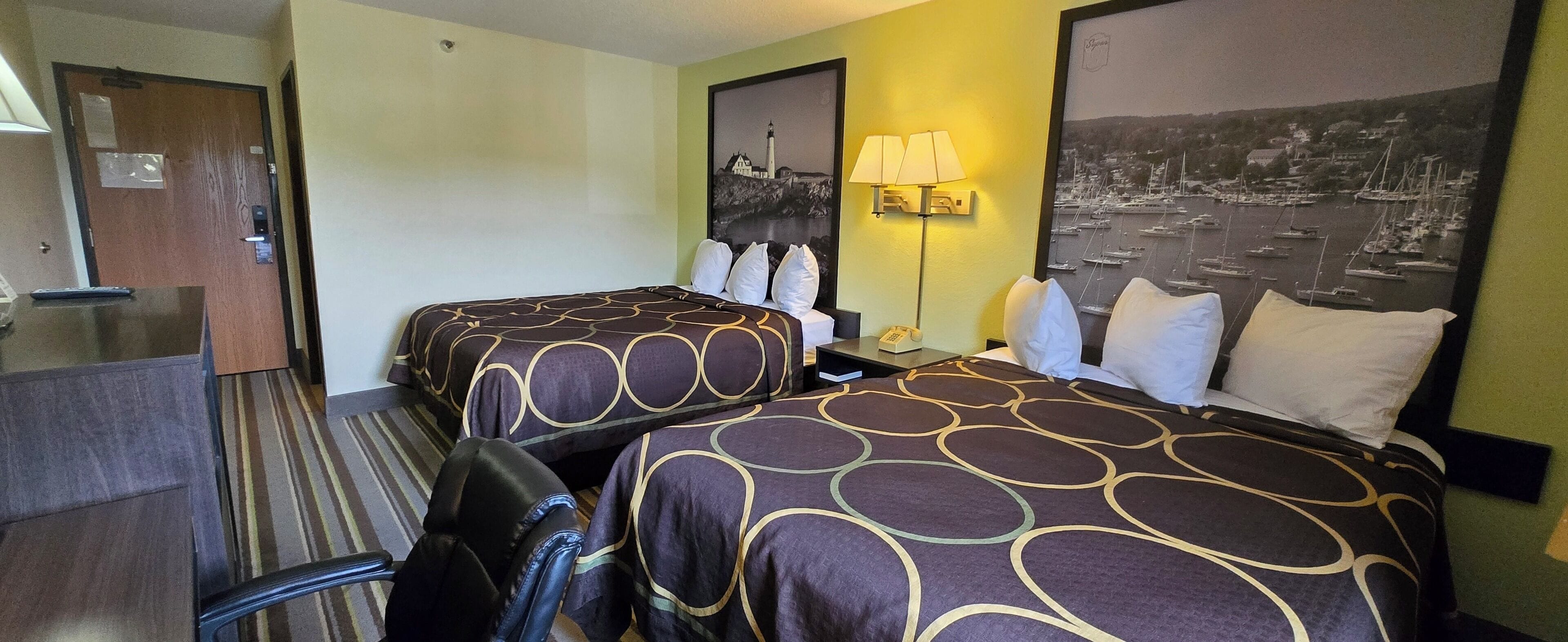 A hotel room with two beds, a desk, a chair, a lamp, and a wall-mounted phone.