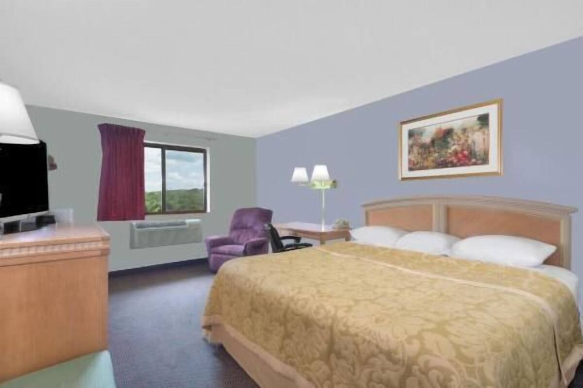 A hotel room with a bed, a desk, a chair, a window with curtains, a painting on the wall, and a lamp.