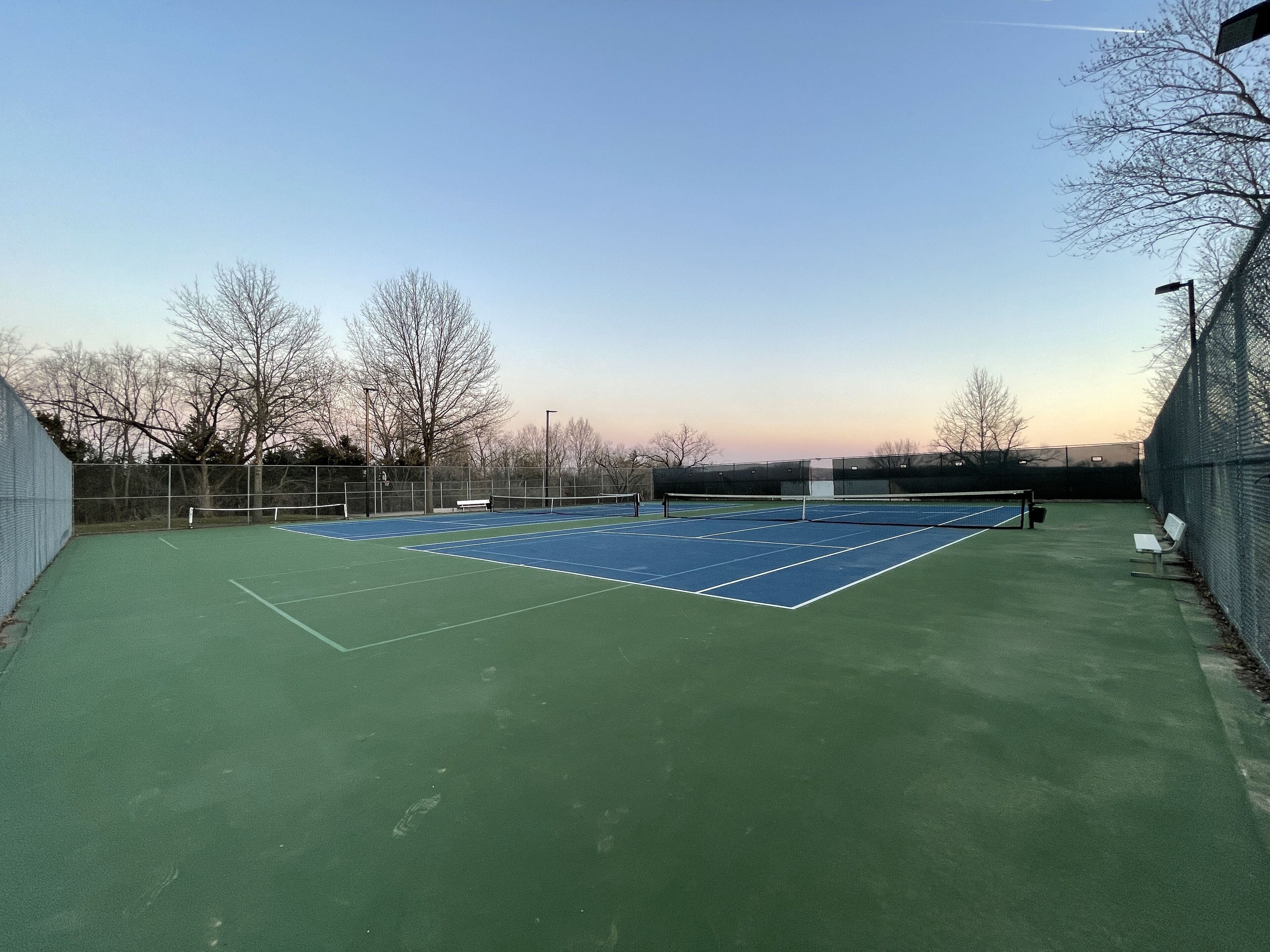 An outdoor tennis court with a net, surrounded by a fence and benches.