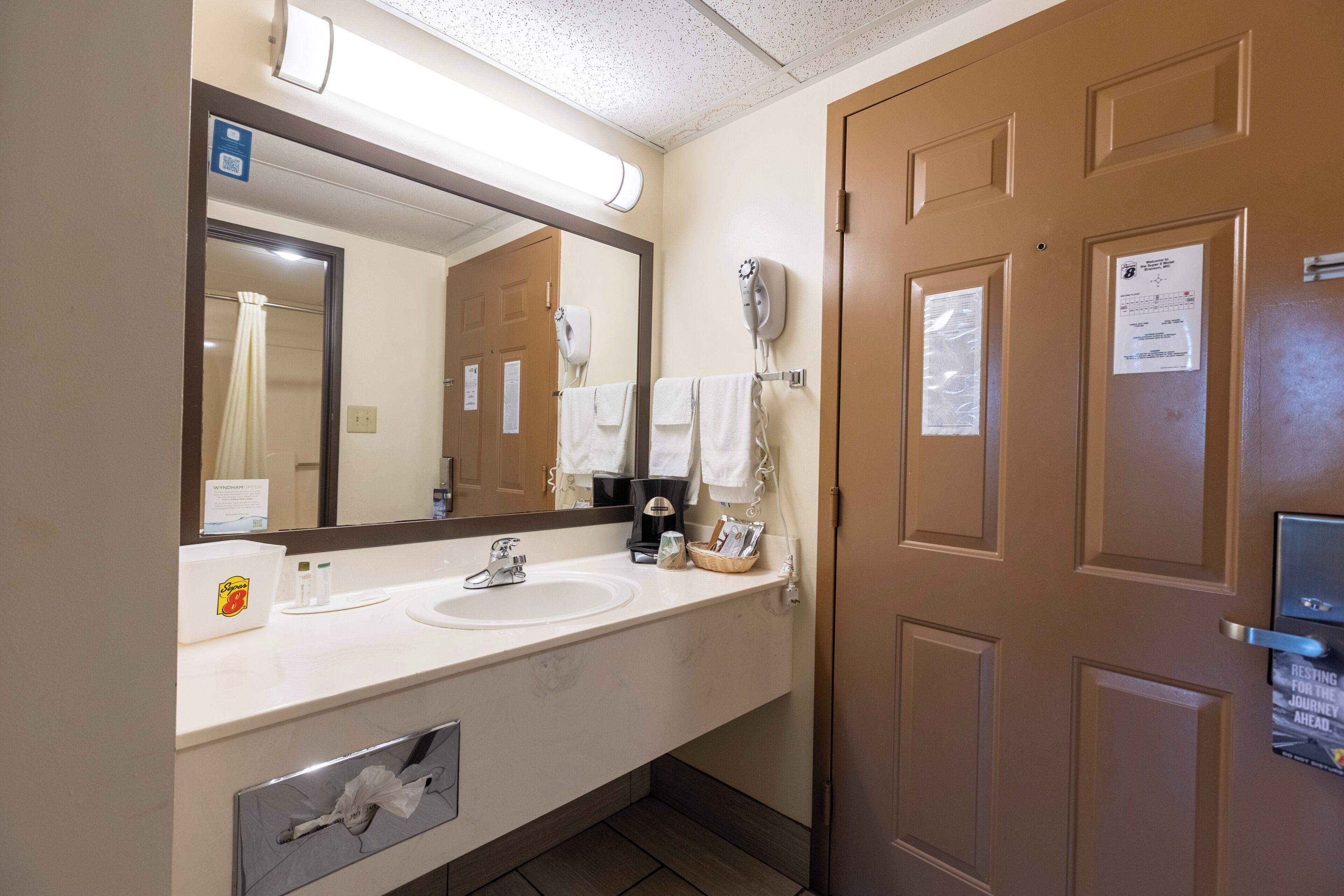 A hotel bathroom with a large mirror, two sinks, a hairdryer, and a door with a sign.