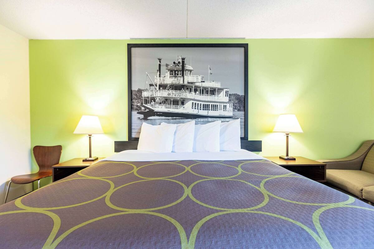 Una habitación de hotel con una cama, dos lámparas de noche, una silla e imagen enmarcada de un barco en la pared.