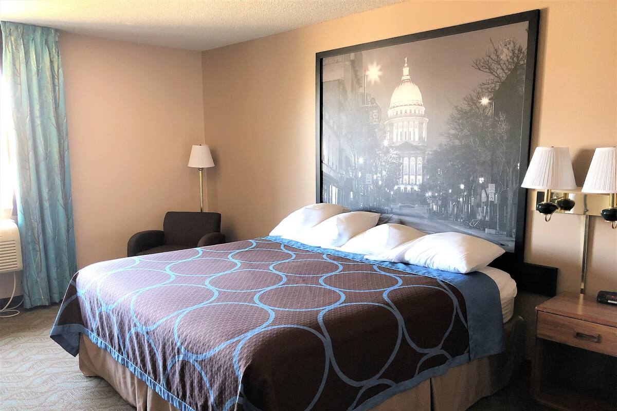 A hotel room with a large bed, a nightstand with two lamps, a chair, a desk, and a large framed picture of a building.