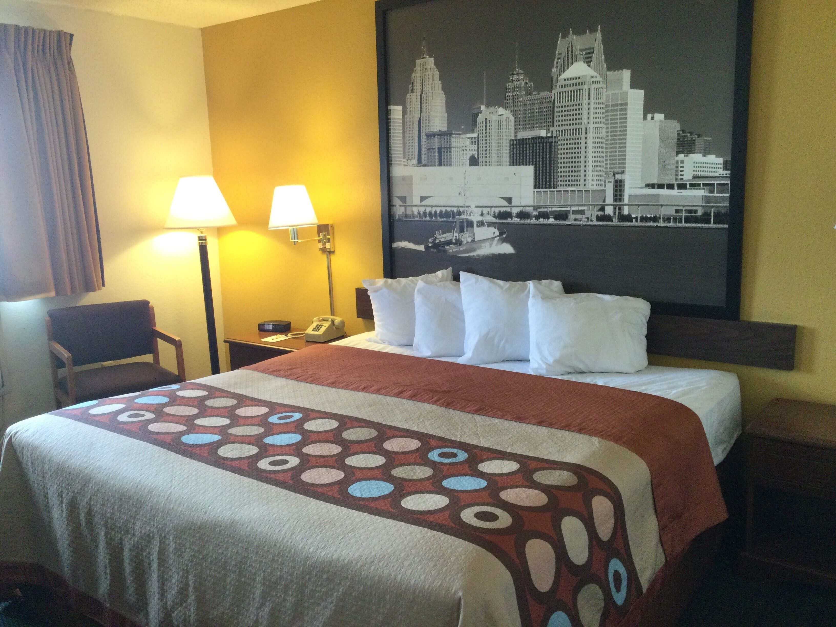 A hotel room with a bed, two bedside lamps, a chair, a nightstand with a phone, and a cityscape painting on the wall.