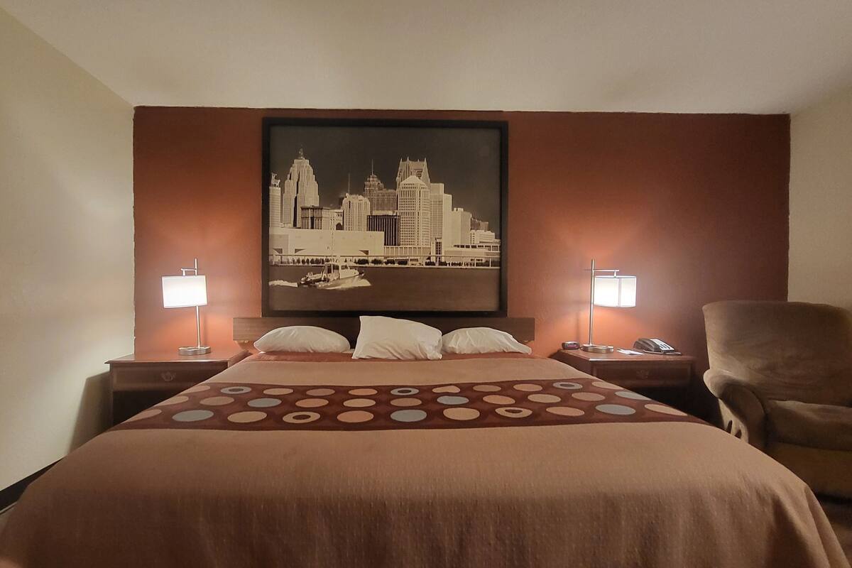 A hotel room with a bed, two bedside lamps, a chair, and a cityscape painting on the wall.