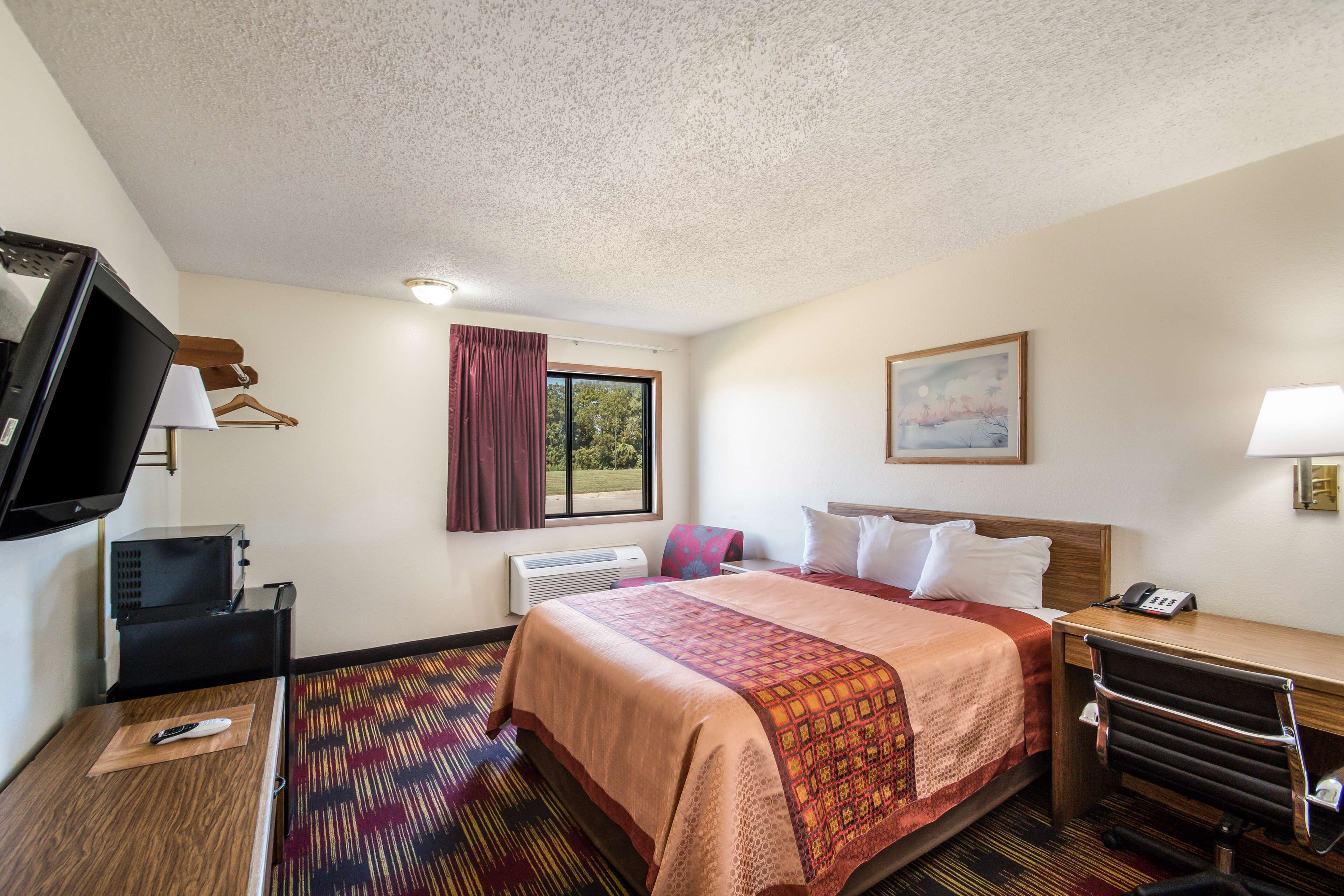 Een hotelkamer met een bed, een bureau, een stoel, een televisie, een verwarming, een raam met gordijnen en een schilderij aan de muur.