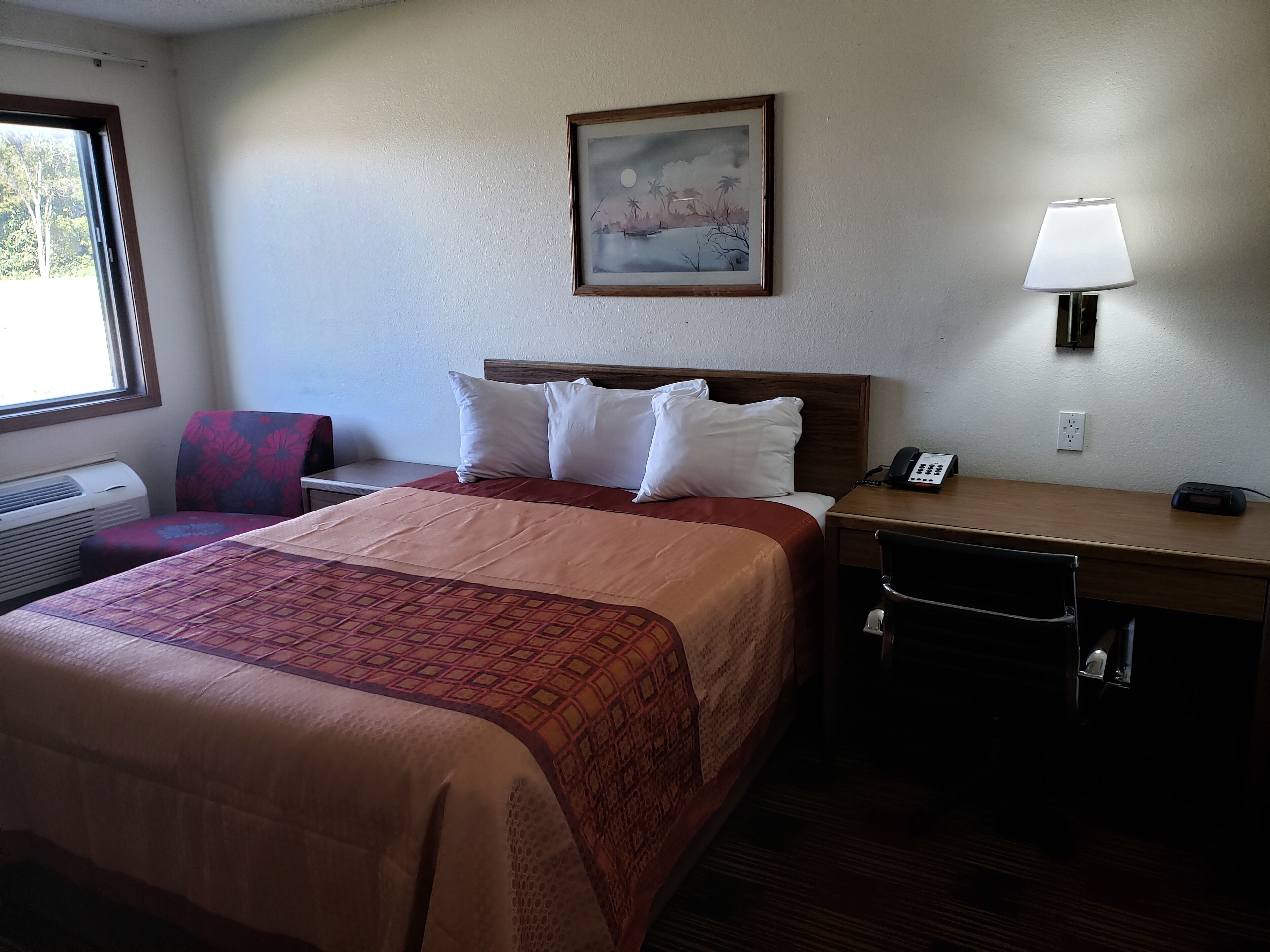 Une chambre d’hôtel avec un lit, un bureau, une chaise, une lampe, un téléphone et un tableau au mur.