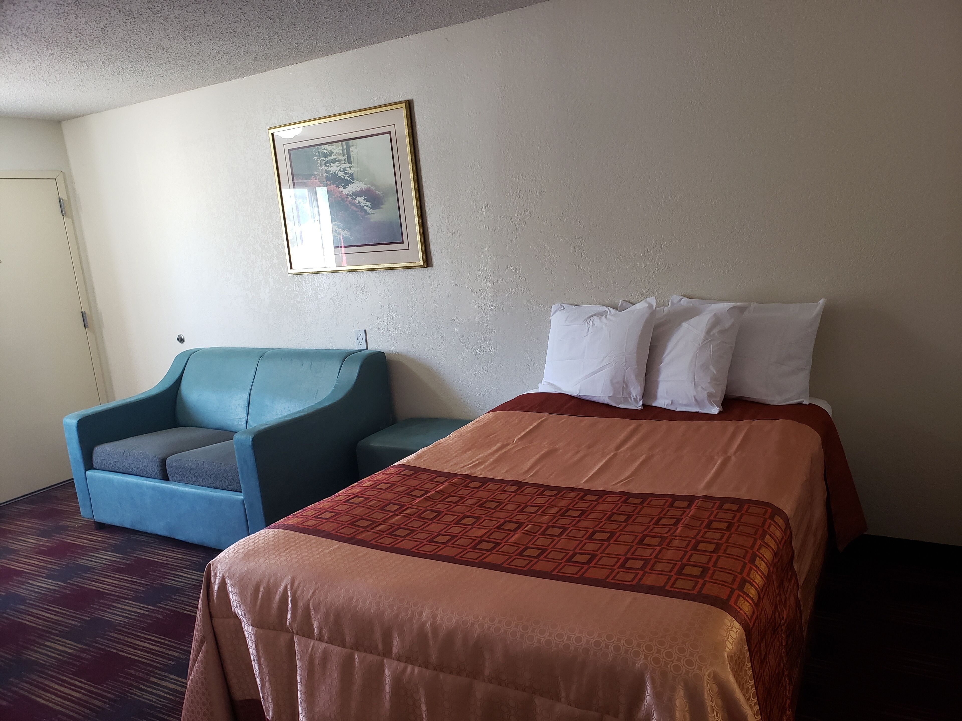 A hotel room with a bed, a blue armchair, and a framed picture on the wall.