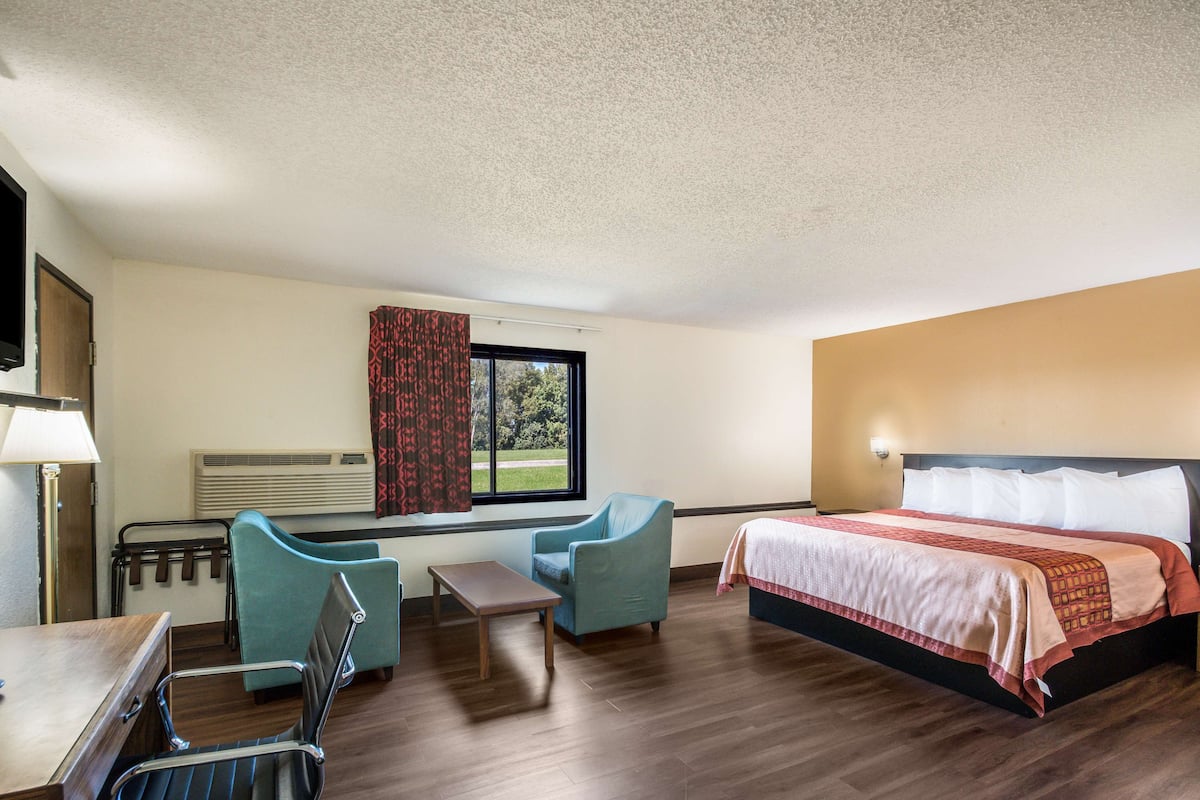 A hotel room with a bed, two chairs, a small table, a window with curtains, and a television on the wall.