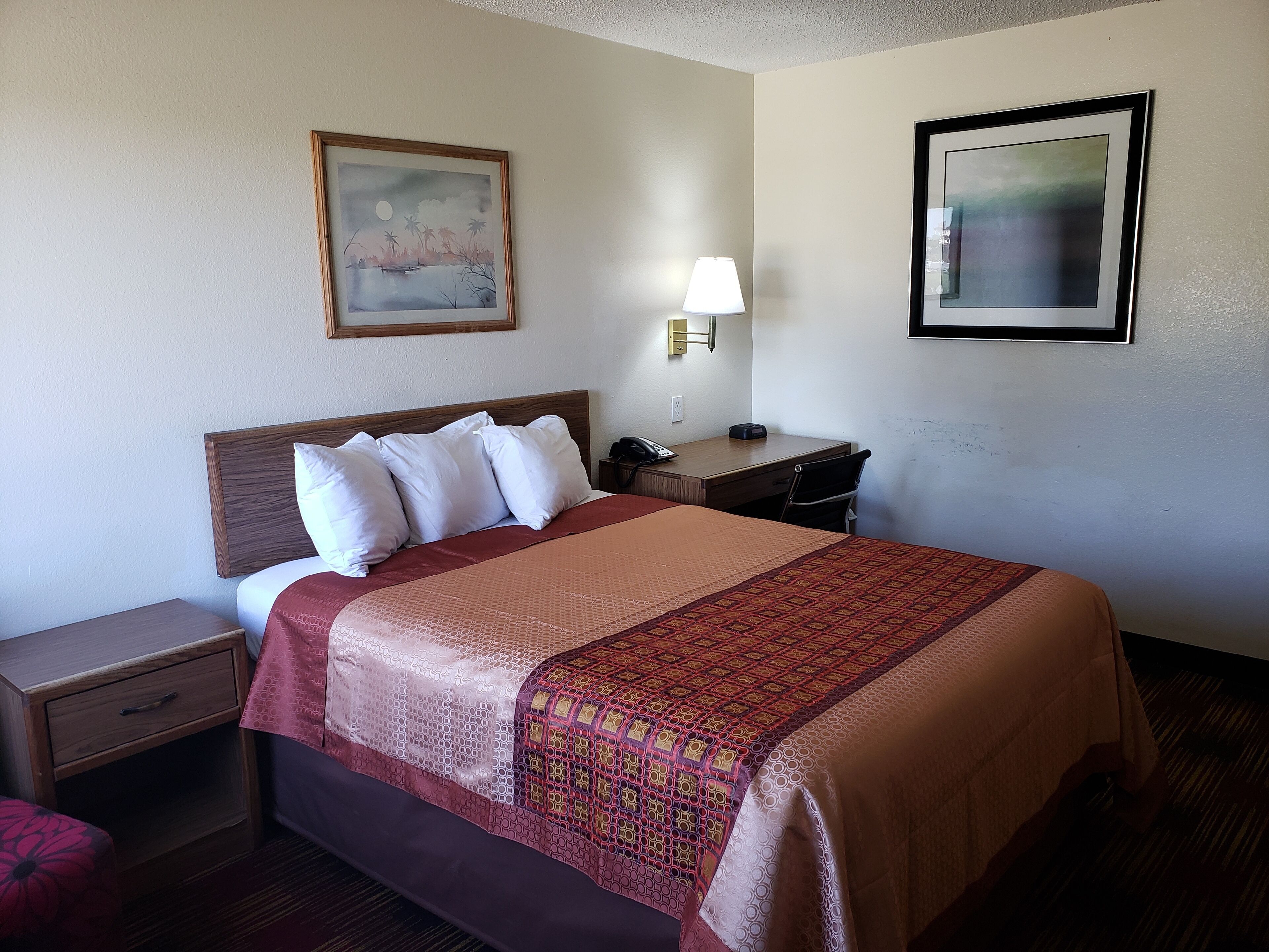 A hotel room with a bed, a nightstand, a desk, a lamp, and two framed pictures on the wall.