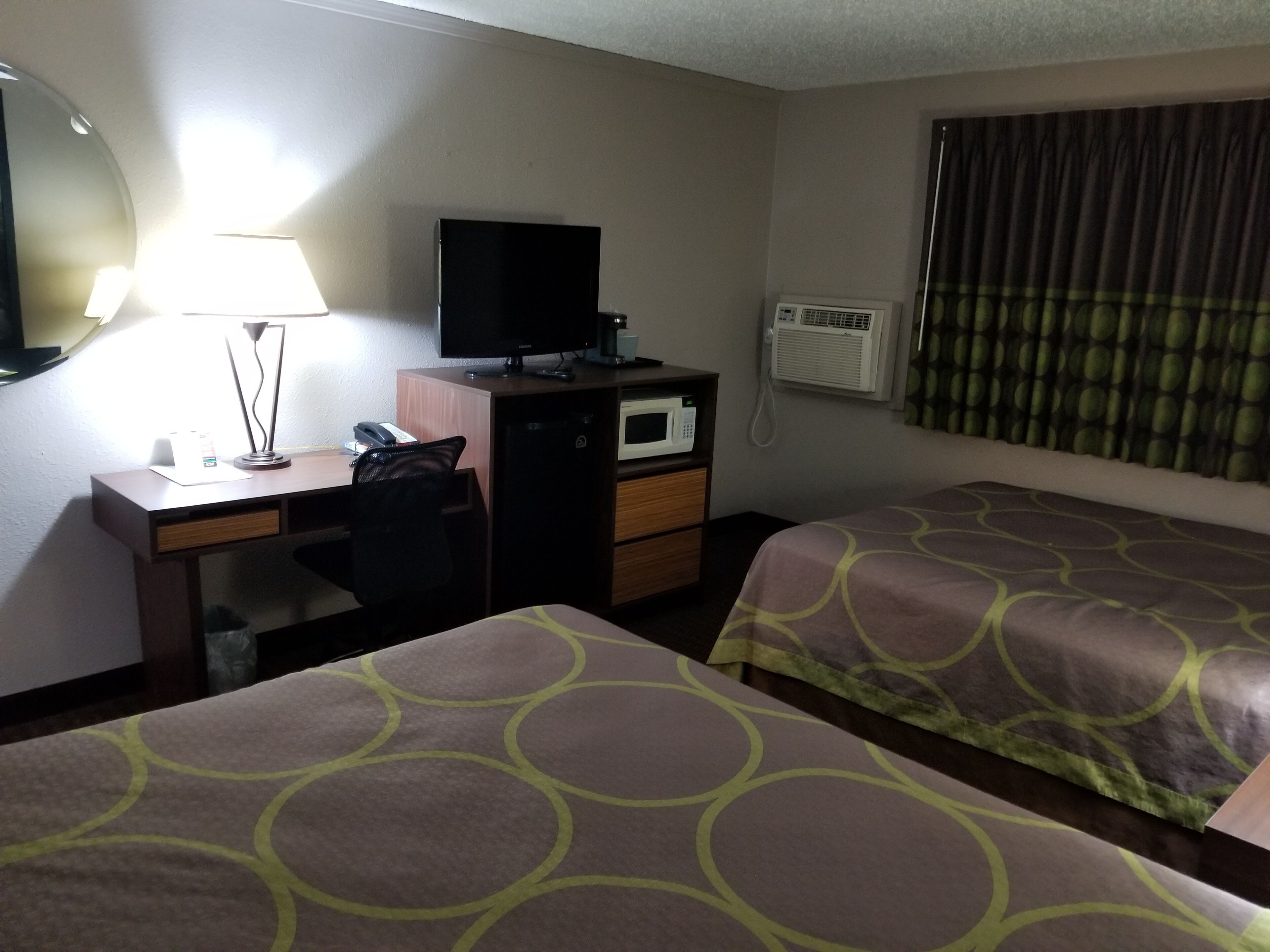 A hotel room with two beds, a desk with a computer, a lamp, a TV, and an air conditioning unit.