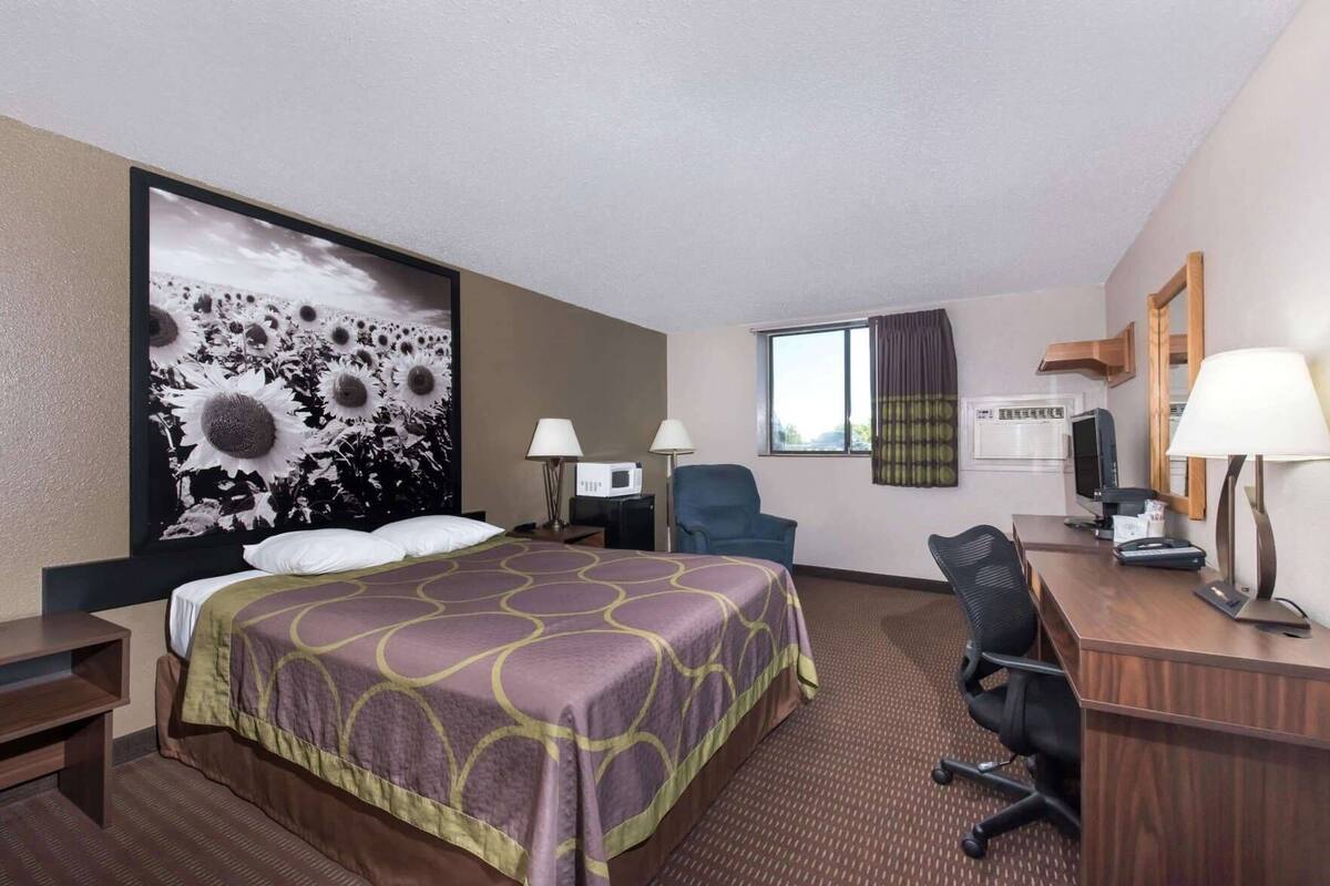 Ein Hotelzimmer mit einem gro ßen Bett, einem Schreibtisch mit Computer, einem Sessel, einem Fernseher und einem großen Wandbild.
