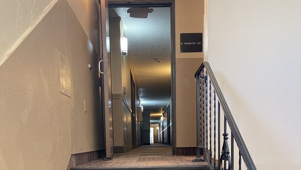 Uma escadaria de madeira com corrimão de metal, levando a um corredor com placas indicando números de quarto.
