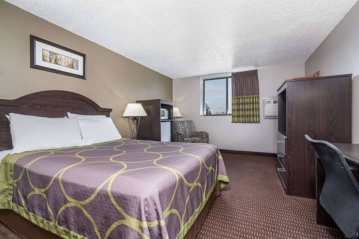 A hotel room with a large bed, a desk, a chair, a lamp, a picture on the wall, and a window with curtains.