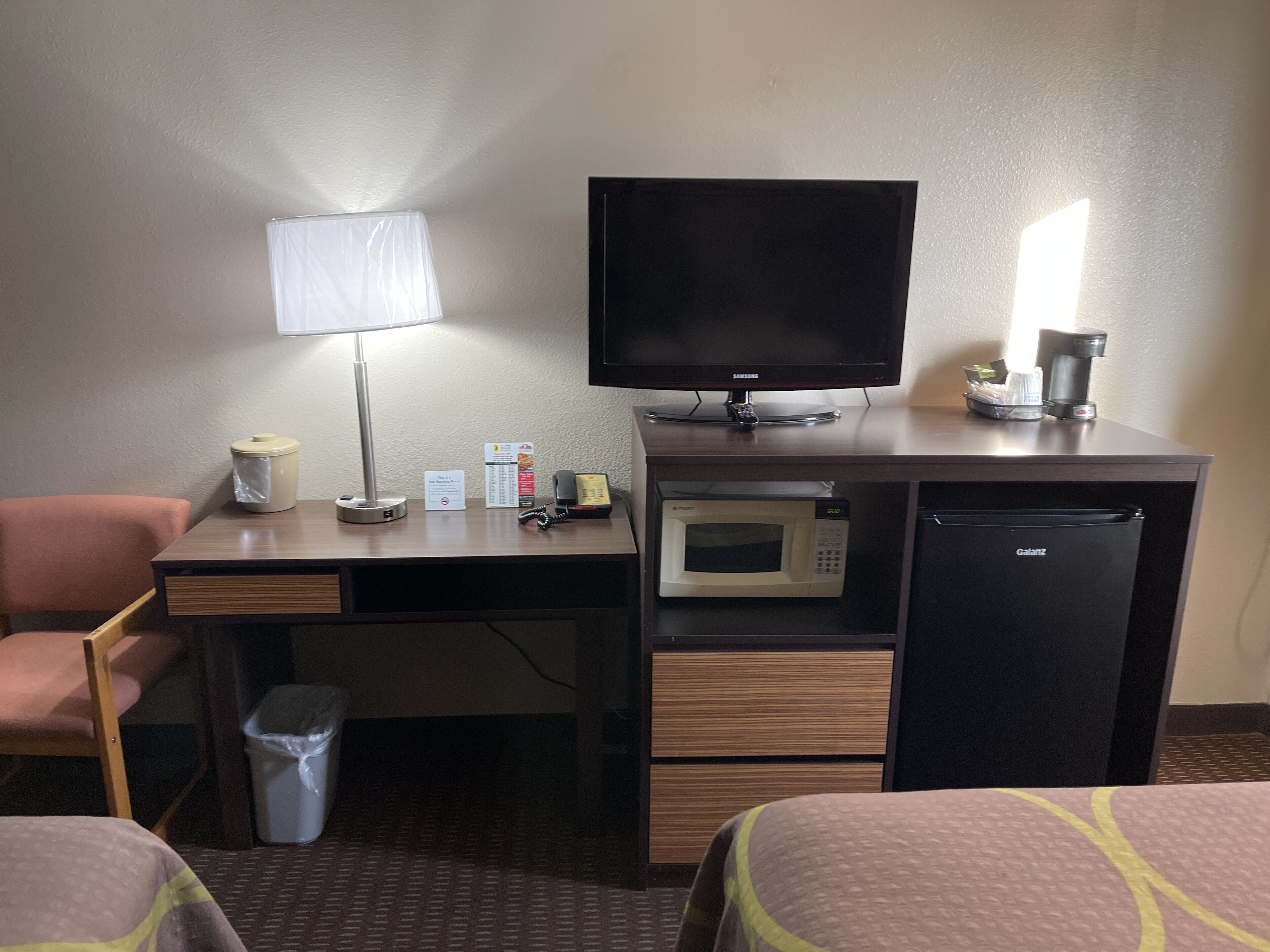 Quarto de hotel com escrivaninha, televisão, micro-ondas, frigorífico, lixeira, candeeiro e uma cadeira.
